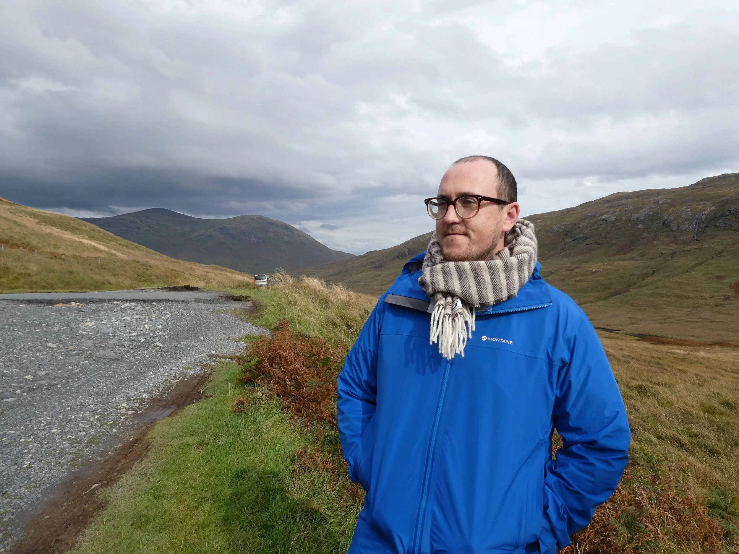 A man wearing glasses, a blue jacket, and a scarf stands outdoors in a mountainous area with gray clouds overhead. There is a small road and a car in the background.