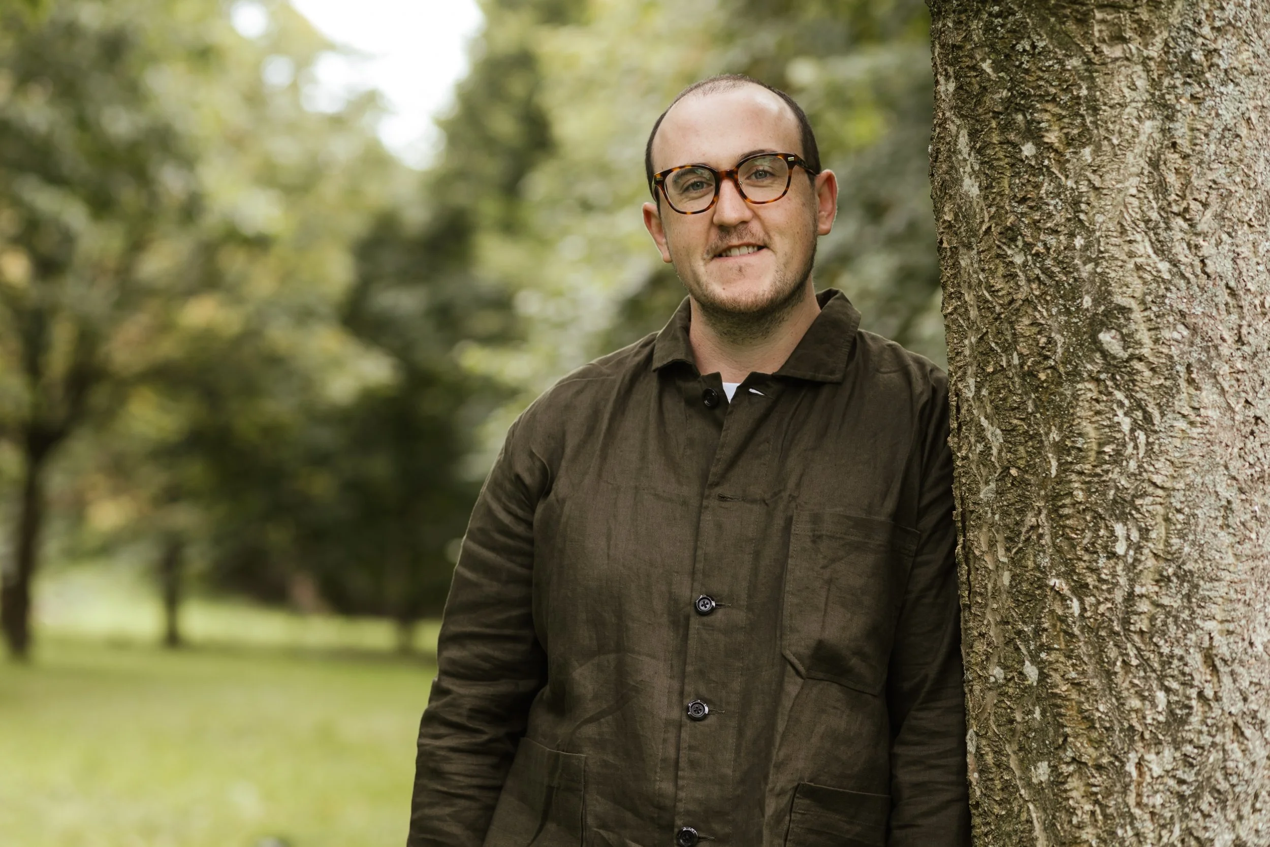 A man wearing glasses and a brown jacket is standing outdoors next to a tree, smiling at the camera with a blurred green background of trees and grass.