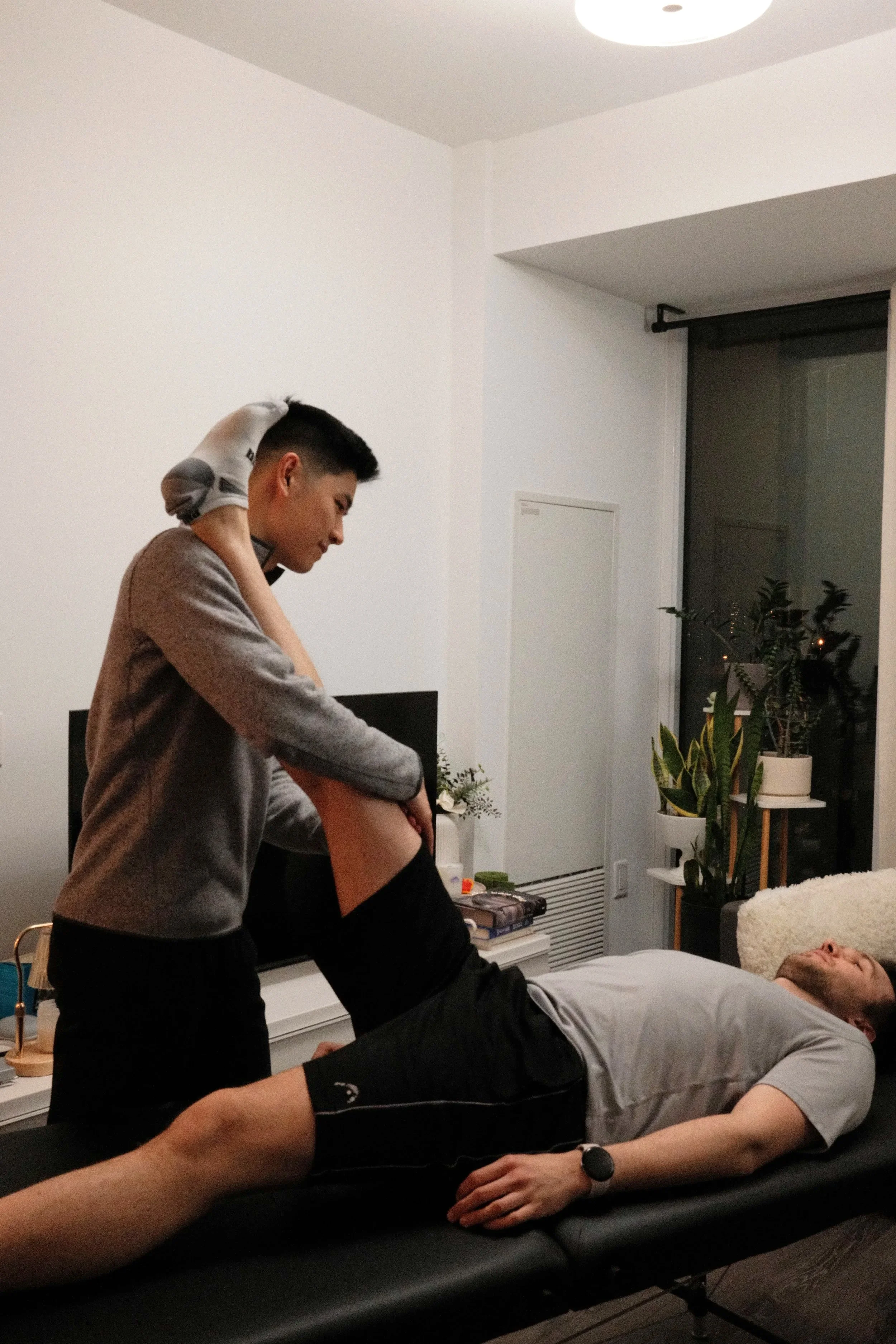 A man lies on a black massage table with his shirt lifted, while a woman stands beside him, massaging his abdomen in a therapy session in a modern living room.