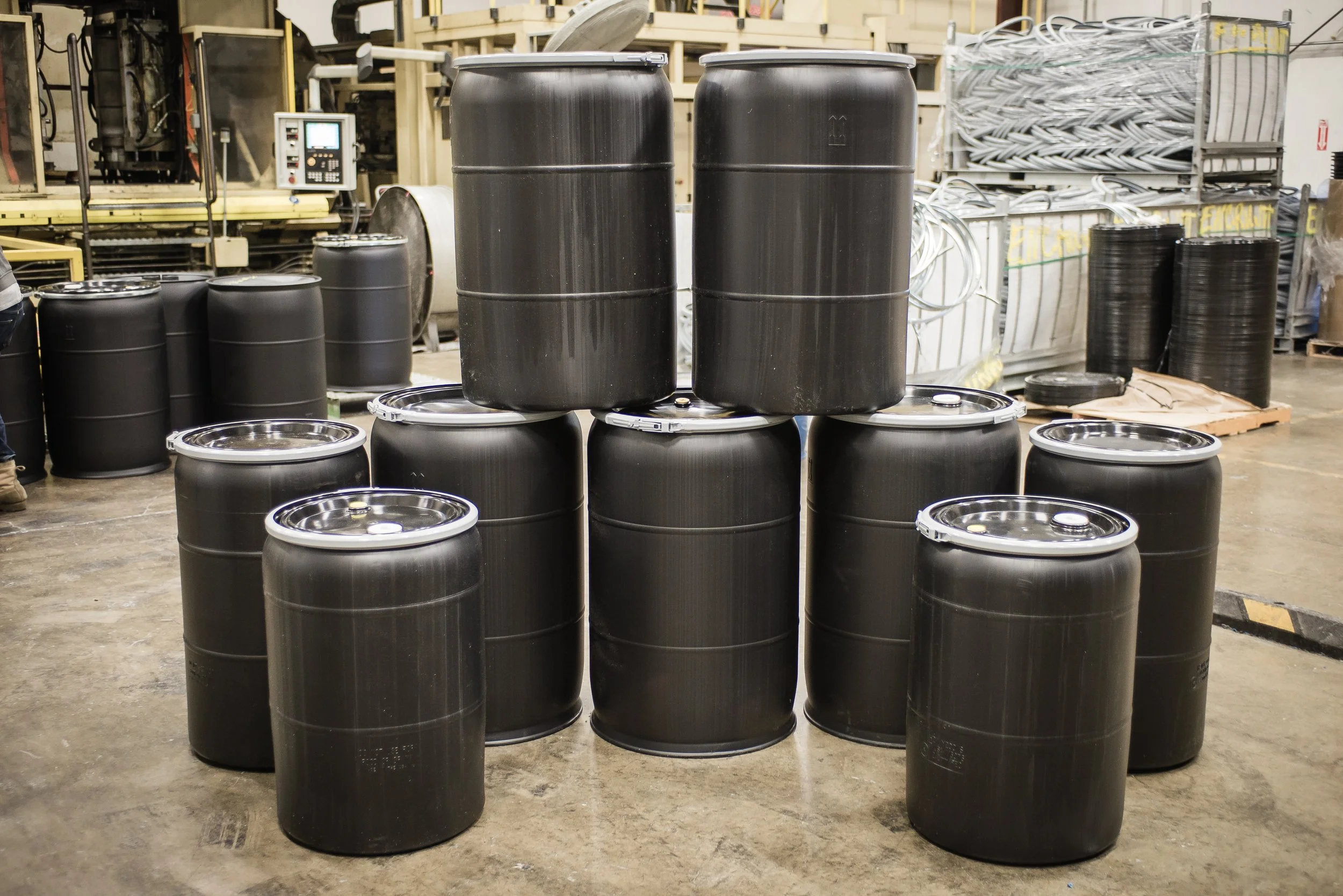 Several black metal barrels with closed lids are arranged in front of industrial machinery and warehouse shelving in a manufacturing or storage facility.