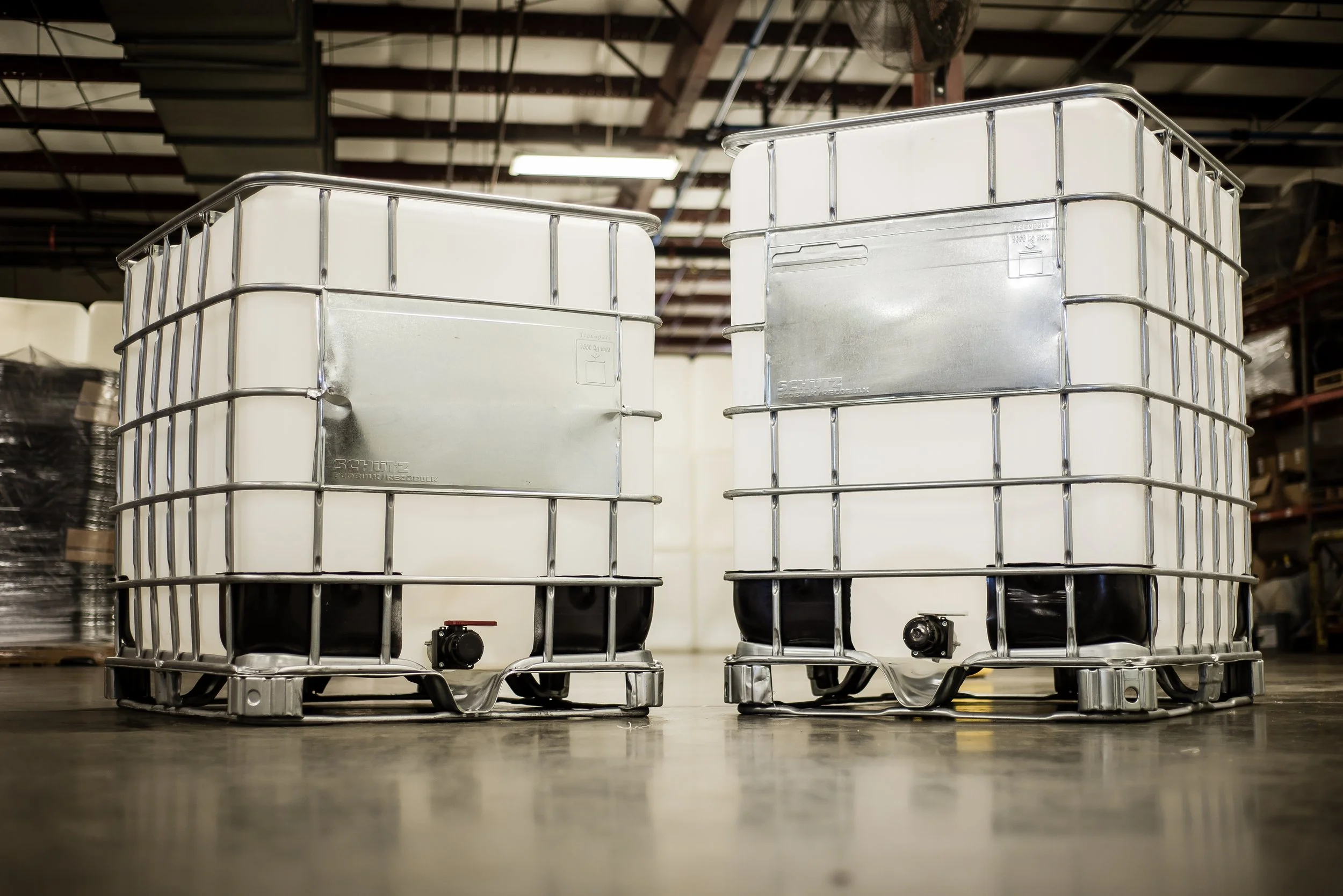Two industrial-grade intermediate bulk containers (IBCs) with white plastic bodies and metal frames, placed on a warehouse floor with shelving and other equipment in the background.