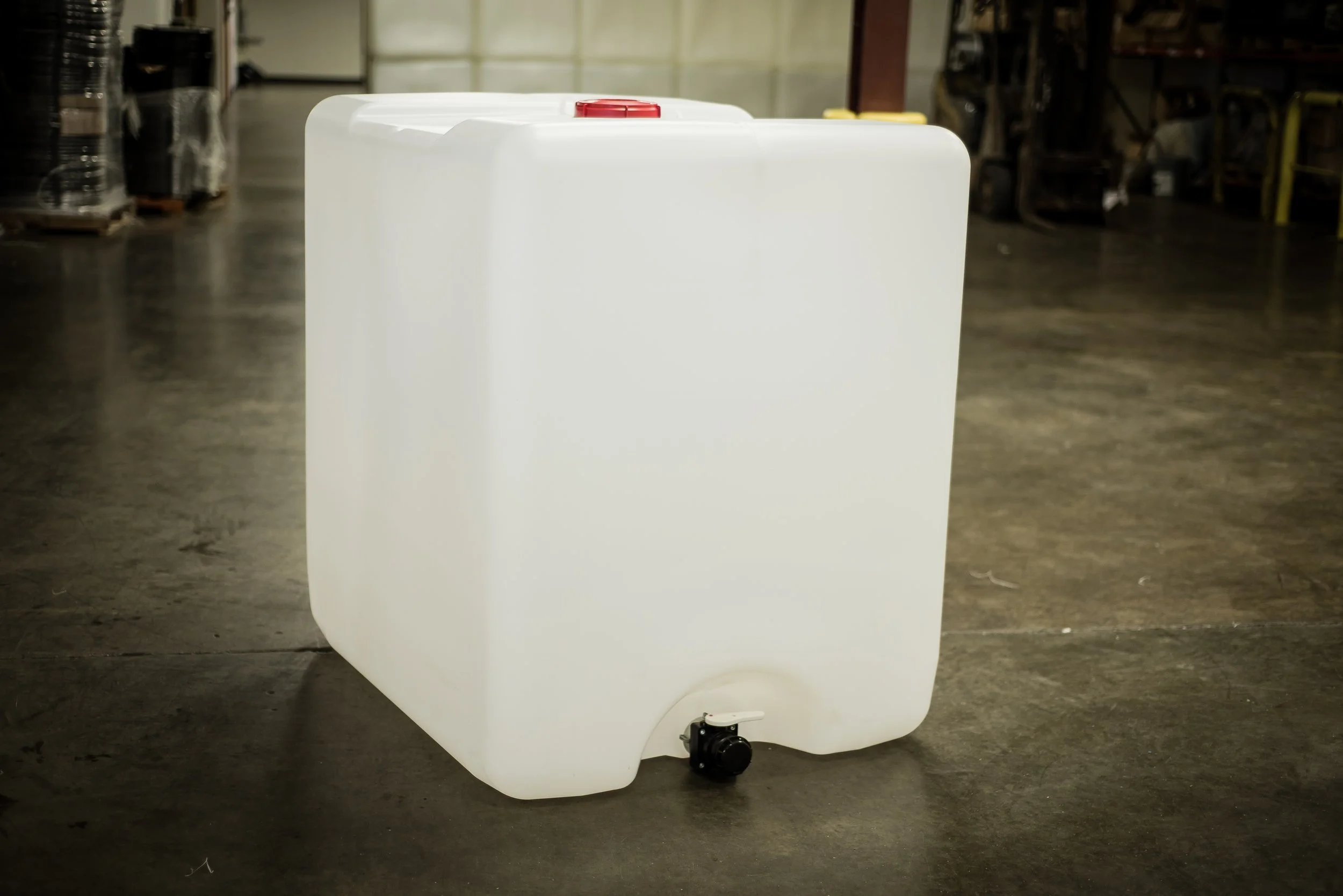 White plastic container with a red cap on top and a spout at the bottom, placed on a concrete floor in an industrial setting.