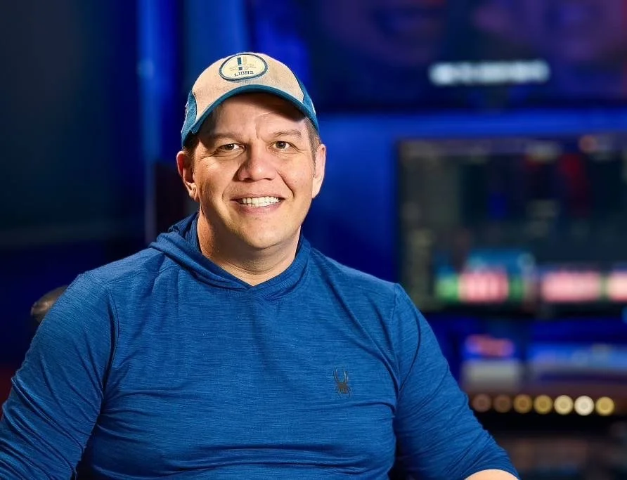 A man wearing a baseball cap and blue hoodie sitting in front of a computer monitor in a dark room.