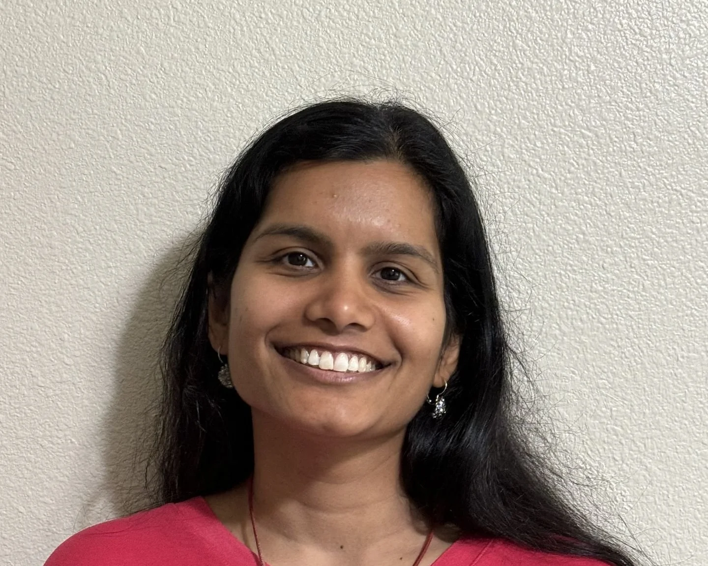 Portrait of a woman with long black hair, smiling, wearing earrings and a pink top, standing against a light-colored textured wall.