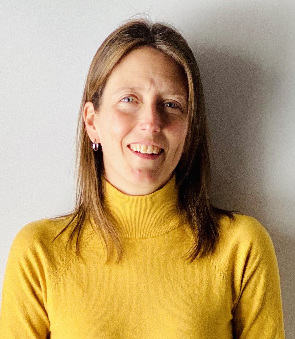 A woman with shoulder-length brown hair, wearing a yellow turtleneck sweater and earrings, smiling at the camera against a plain light-colored background.