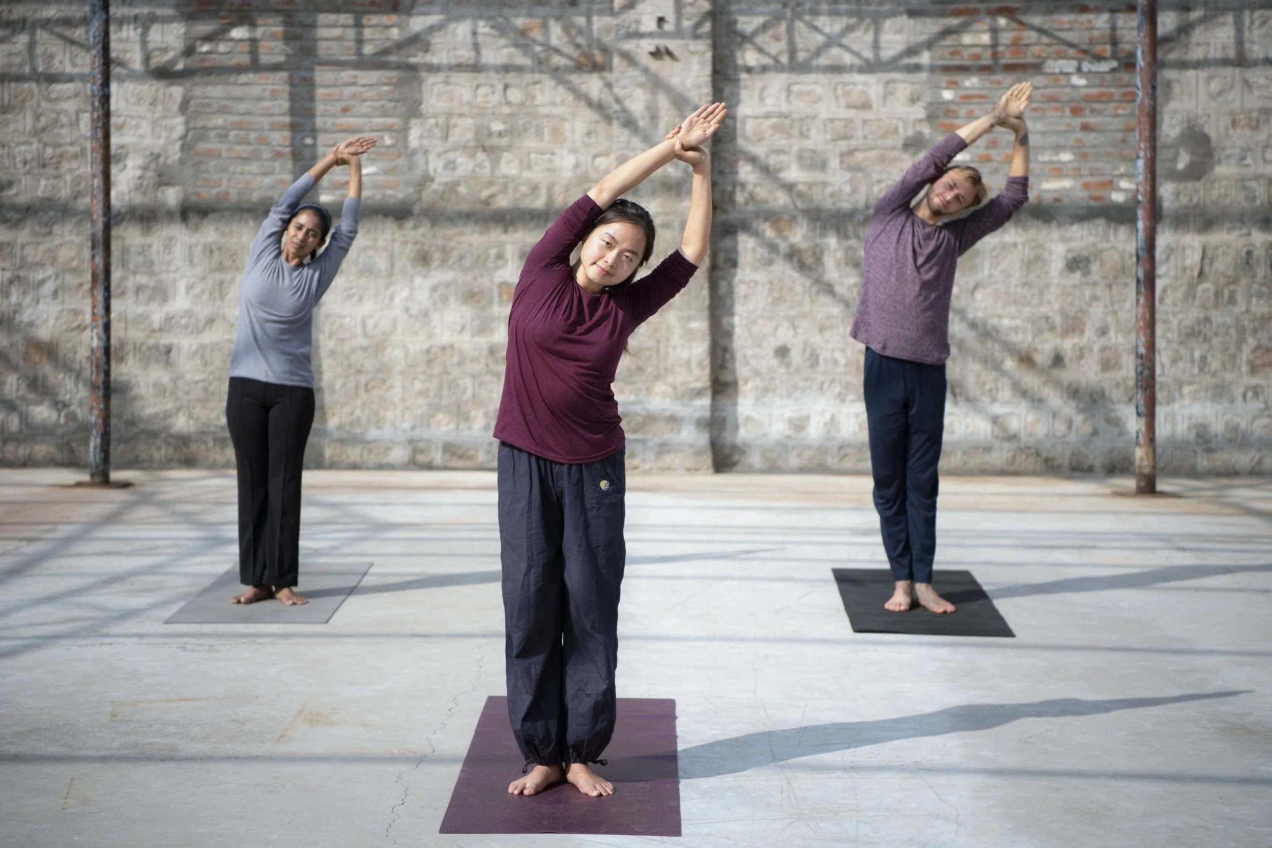 Three women practicing yoga outdoors on mats, engaging in stretching poses against a brick wall background.