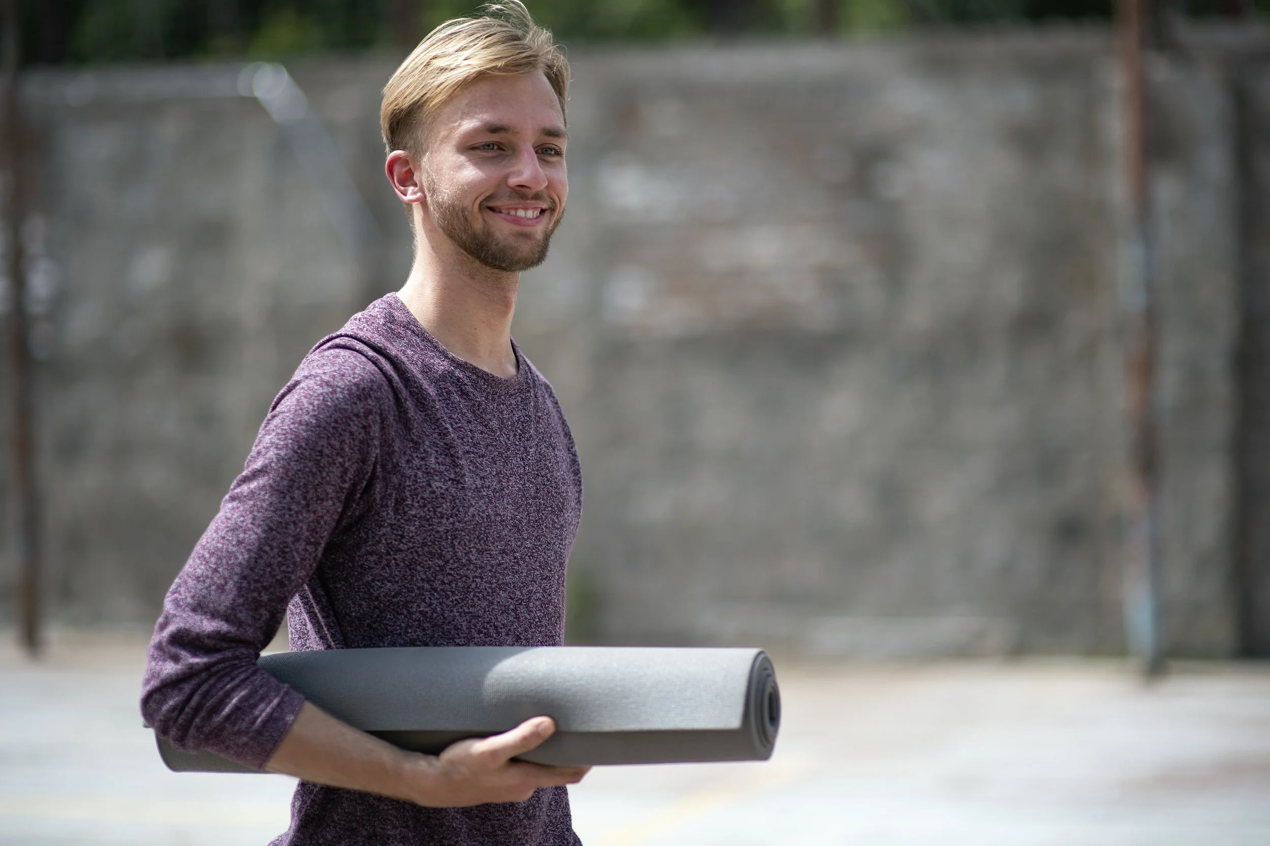Young man holding a rolled yoga mat outdoors, smiling, with an outdoor concrete wall in the background.