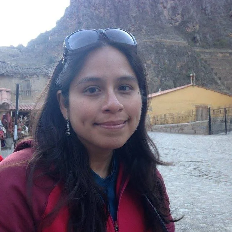 A woman with glasses on her head and earrings, wearing a red jacket, standing outdoors in front of a mountain and a yellow building.