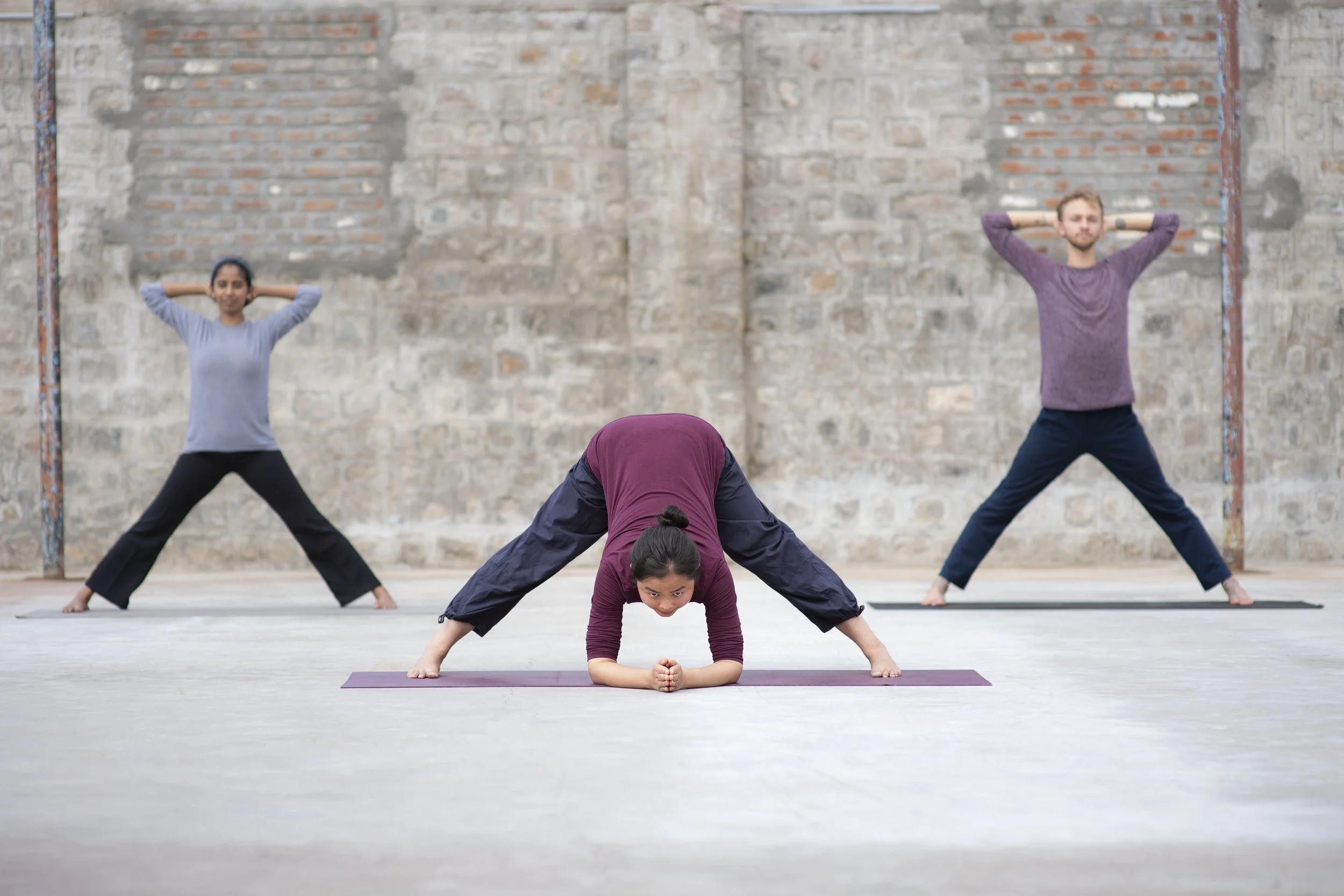 Three people practicing yoga outdoors on mats, with a brick wall in the background. One person in the foreground is in a plank position, while two others in the background are standing with hands behind their heads.