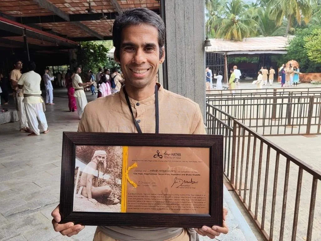 A man smiling and holding a framed certificate at a cultural or religious event with people and animals in the background.