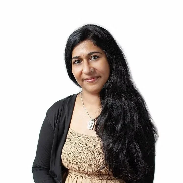 A woman with long black hair, wearing a beige top, black jacket, and a silver necklace with a pendant, smiling against a plain white background.