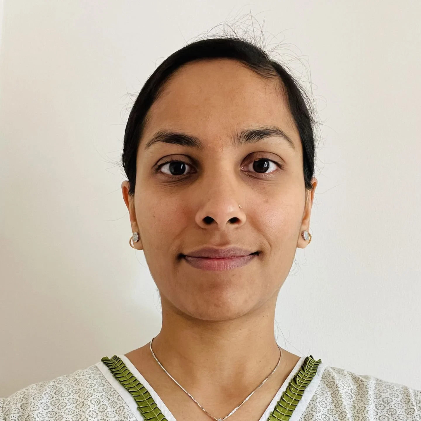 Close-up portrait of a woman with dark hair and earrings, smiling softly, wearing a white top with green trim, against a plain light-colored background.