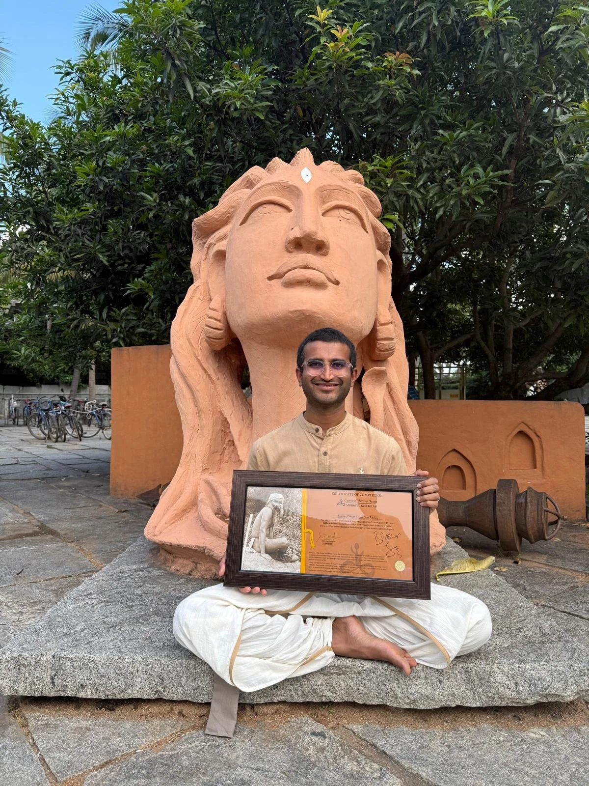 Man dressed in traditional Indian attire sitting in a cross-legged position on a stone platform, holding a framed certificate, with a large clay sculpture of a woman’s face and headdress behind him, and trees in the background.