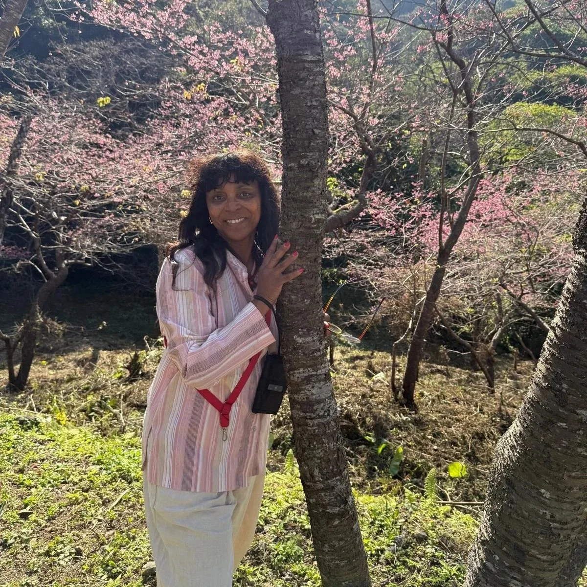 A woman in a pink and white striped blazer and off-white pants standing next to a tree with pink blossoms in a park, smiling at the camera.