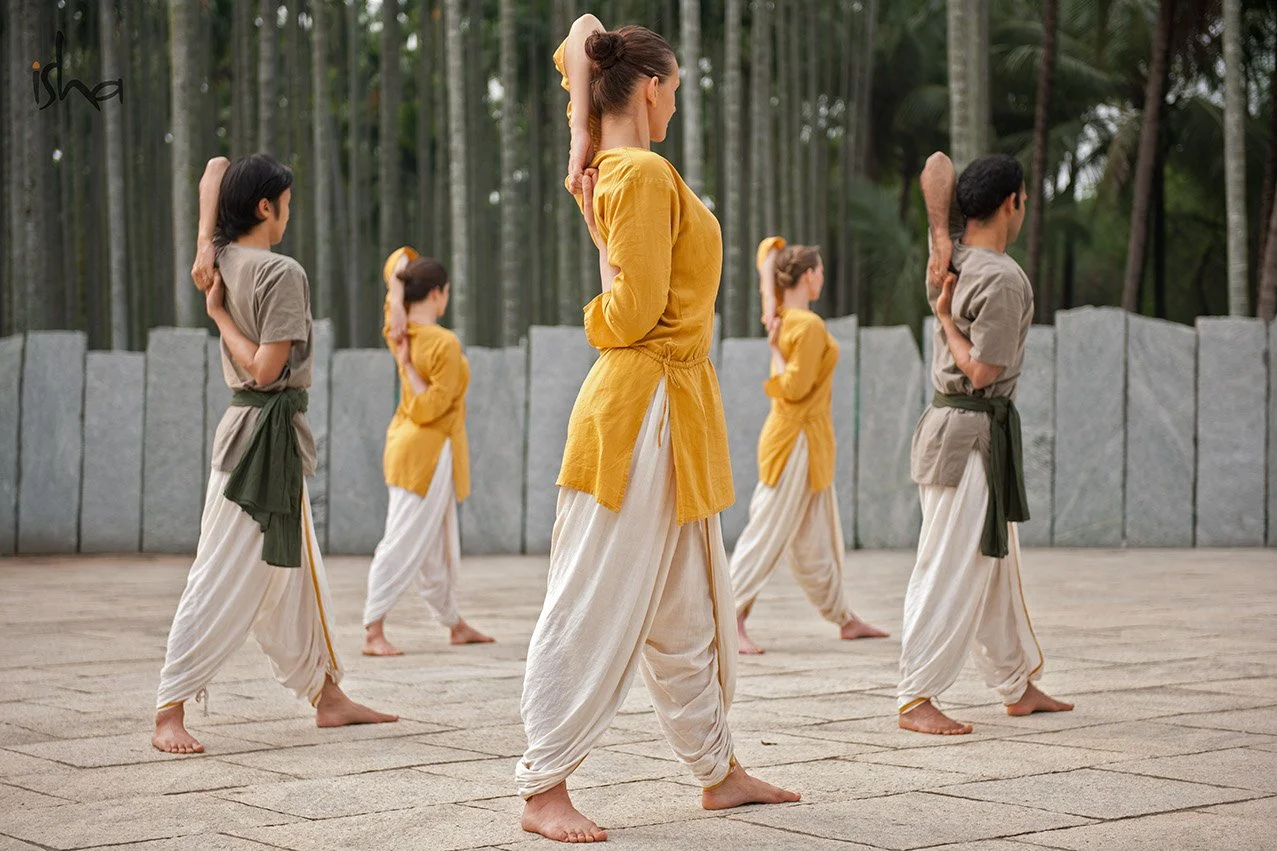 A group of six women practicing yoga outdoors, standing in a row with right arm bent behind their heads and left arm holding the right elbow, in a serene natural setting with trees in the background.