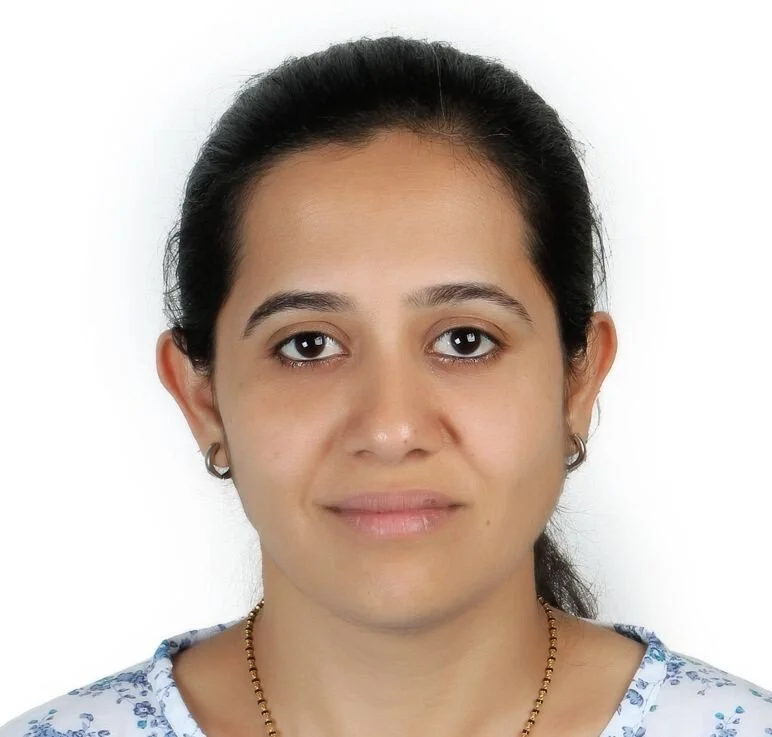 A woman with dark hair pulled back, wearing a white top with blue floral pattern, small hoop earrings, and a gold necklace, looking directly at the camera against a plain white background.