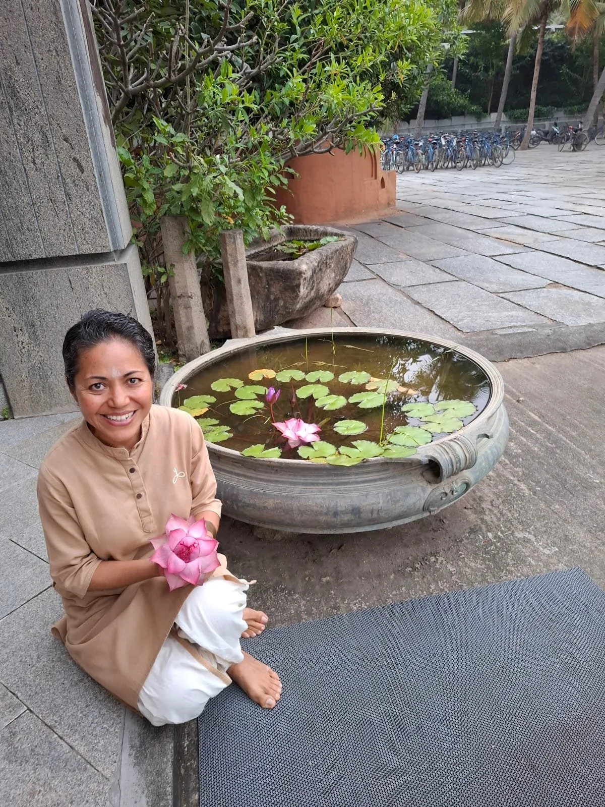 A woman sitting on the ground holding a pink lotus flower next to a large round water basin with lotus flowers and green leaves, outdoors near a building and a parking lot with bicycles.