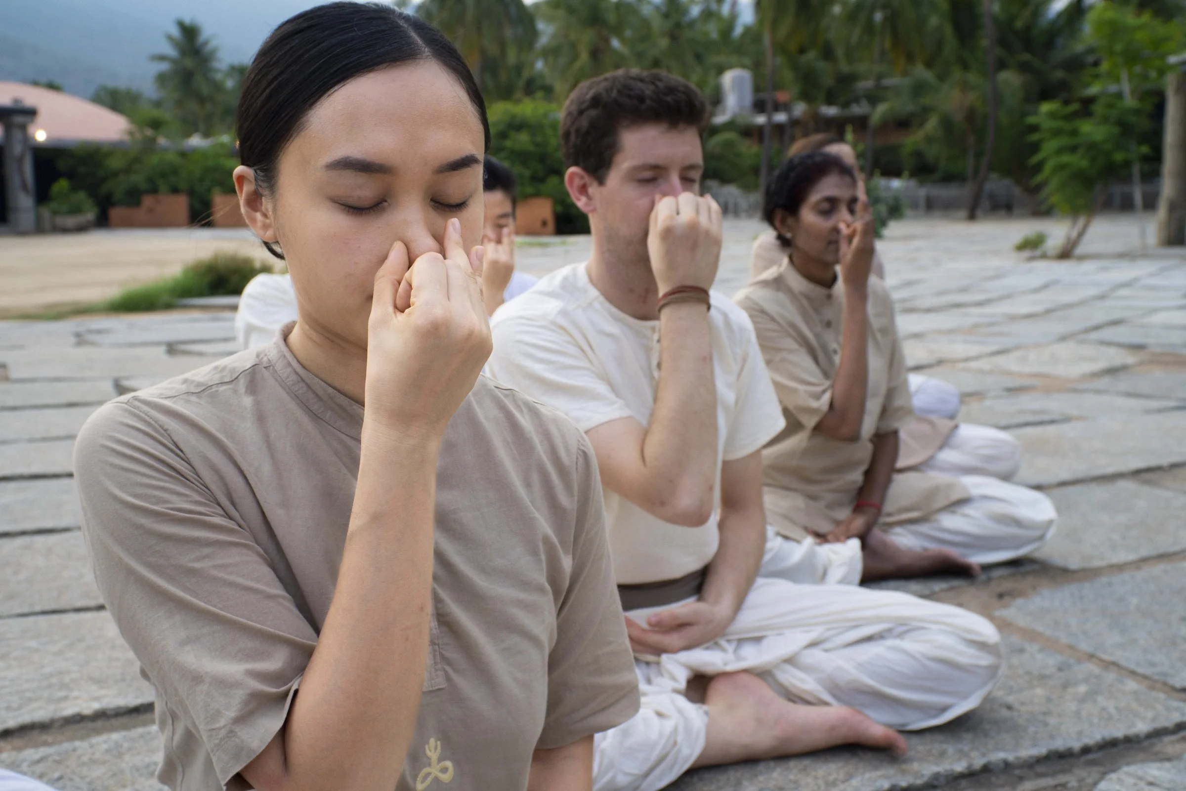Group of people practicing meditation outdoors, sitting cross-legged on a stone surface with eyes closed and hands in a mudra pose