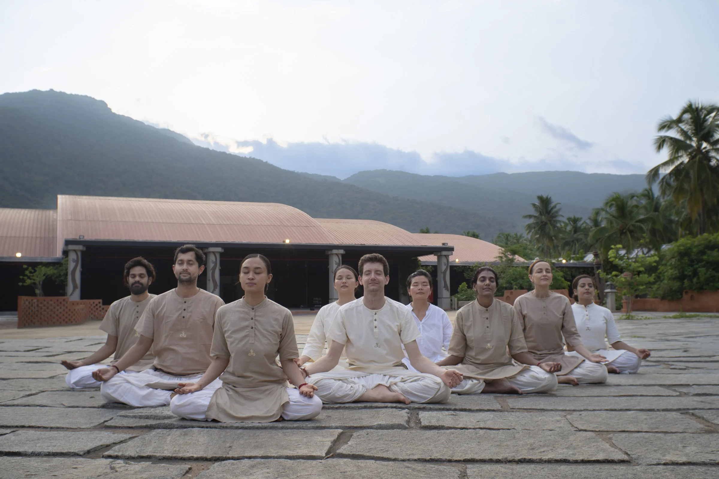 Ten people practicing meditation outdoors on a stone patio, seated in a cross-legged position with closed eyes, with mountains and palm trees in the background.