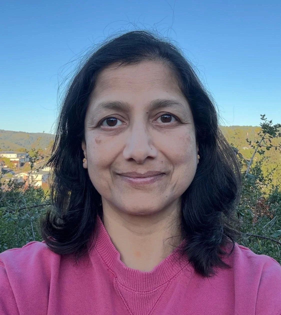 A woman with black hair, wearing a pink top, standing outdoors with a clear sky and distant hills in the background.