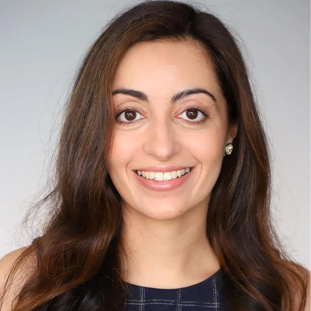 Close-up portrait of a young woman with long brown wavy hair, wearing pearl earrings, smiling, in a dark plaid top, against a light gray background.