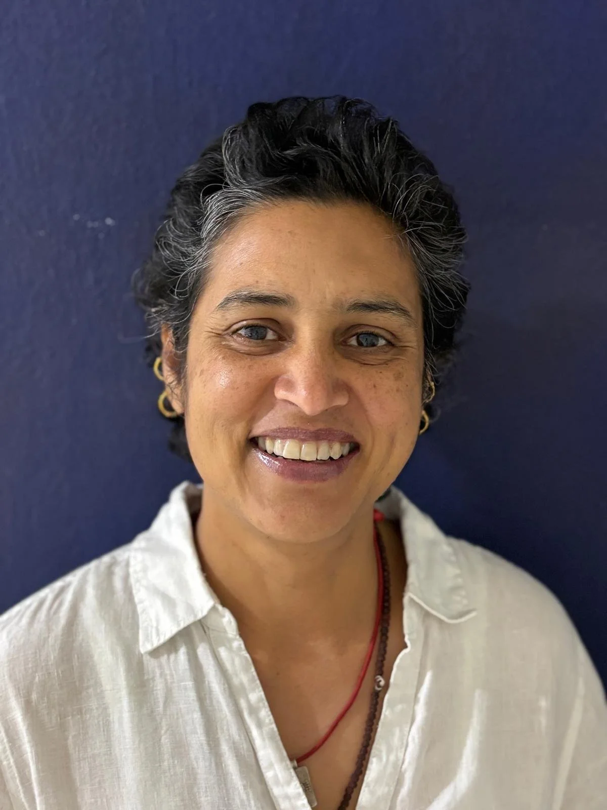 Close-up of a woman with short black hair, light brown skin, and blue eyes, smiling against a dark blue wall. She is wearing a cream-colored shirt, gold hoop earrings, and red and black necklaces.