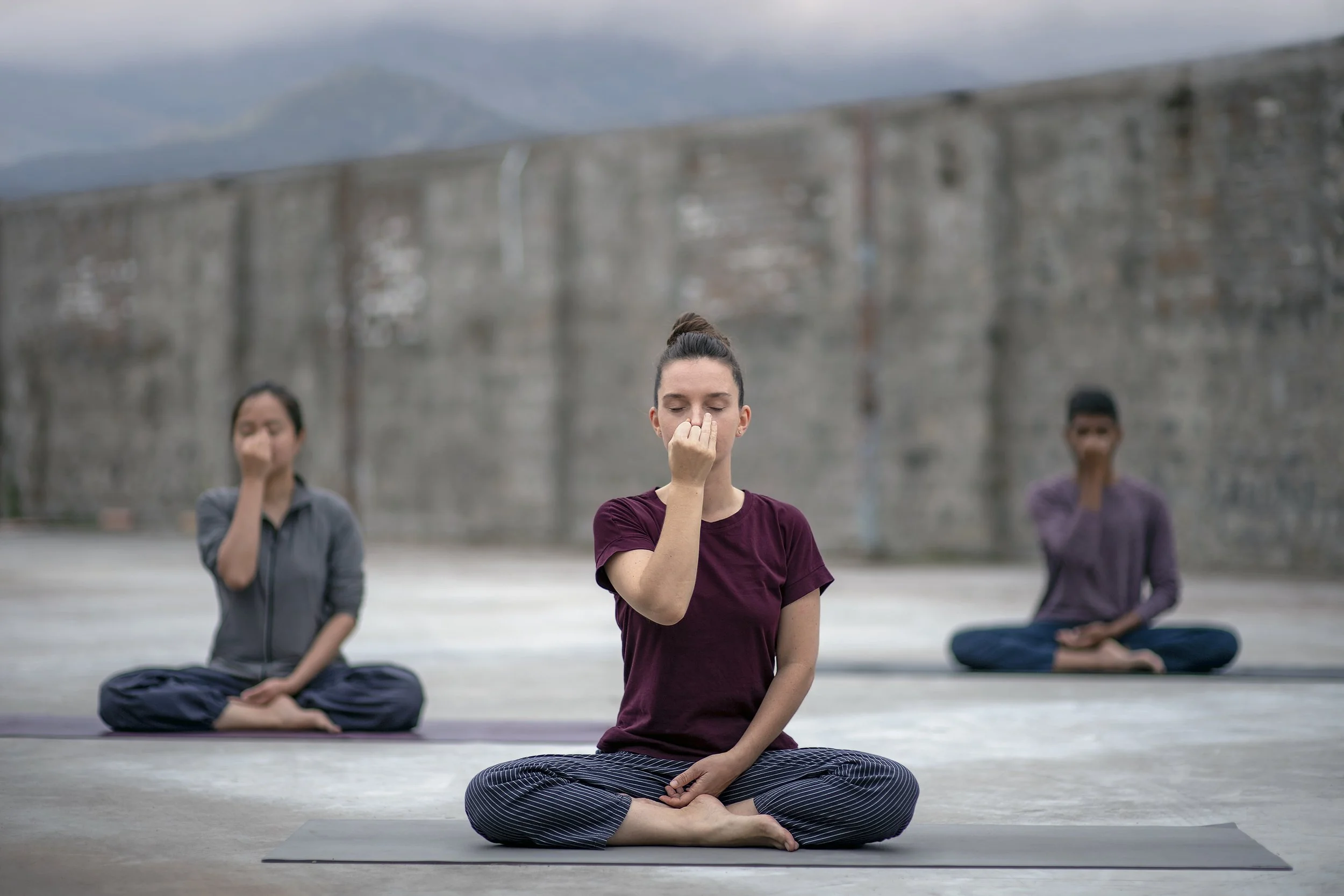 Three women practicing yoga outdoors on gray mats in front of a concrete wall, with mountains in the background. They are sitting cross-legged, with their eyes closed, and both hands covering their nose in a meditative pose.