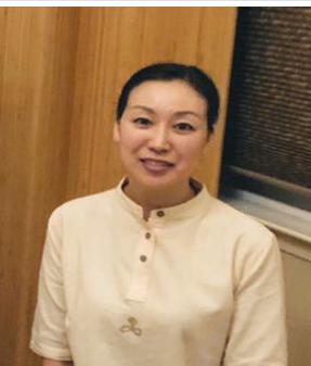 A woman with dark hair pulled back, wearing a cream-colored collared shirt, standing indoors against a wooden wall with a window.