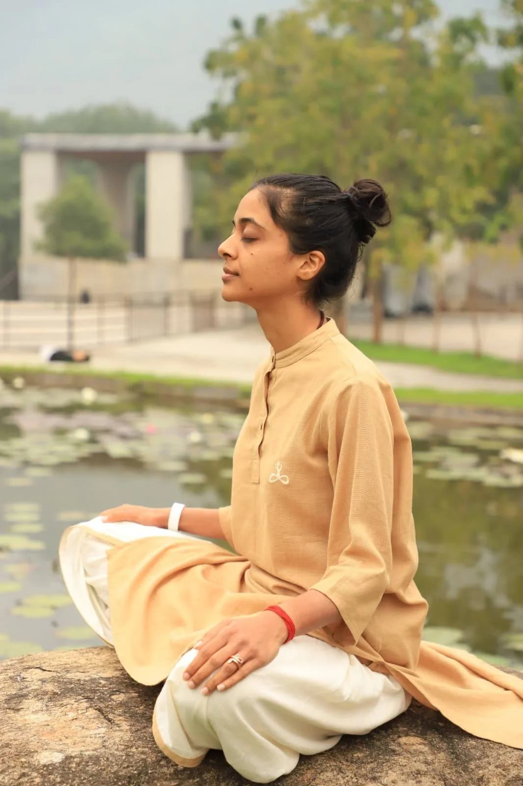 A woman sitting cross-legged on a rock beside a pond with lily pads, practicing meditation with her eyes closed, wearing a beige kurta and white pants, with trees and a modern building in the background.