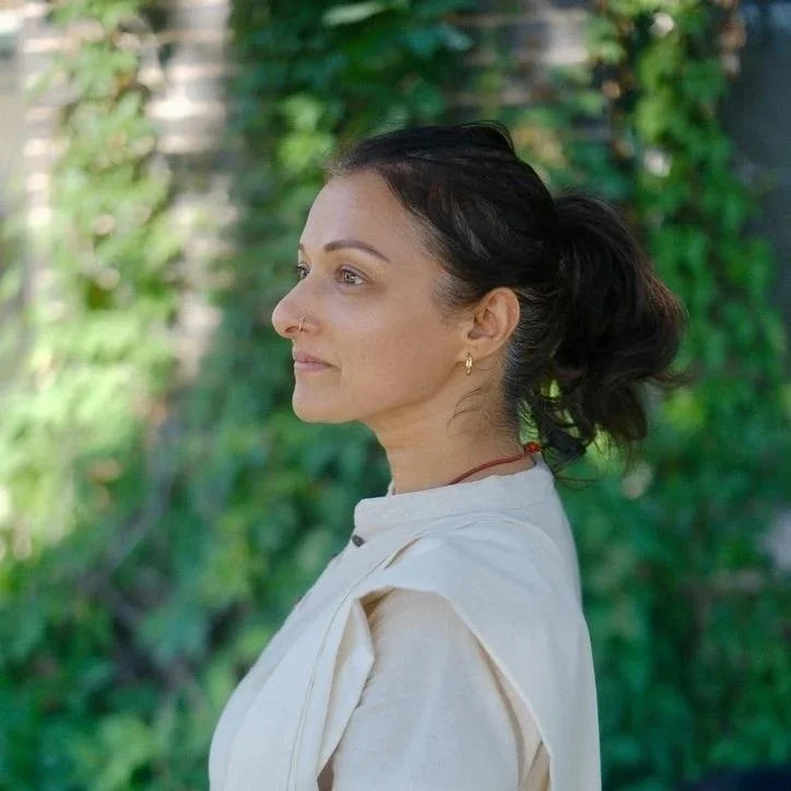 Side view of a woman with dark hair tied back, wearing a white jacket, standing outdoors with a green leafy background.