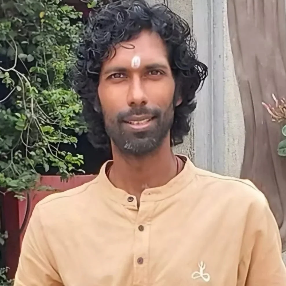 A man with curly black hair and a beard, wearing a beige kurta, standing outdoors with greenery and a wall in the background.