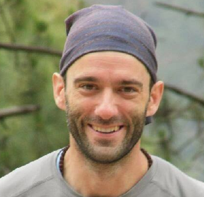 Man smiling outdoors wearing a headscarf with trees in the background.