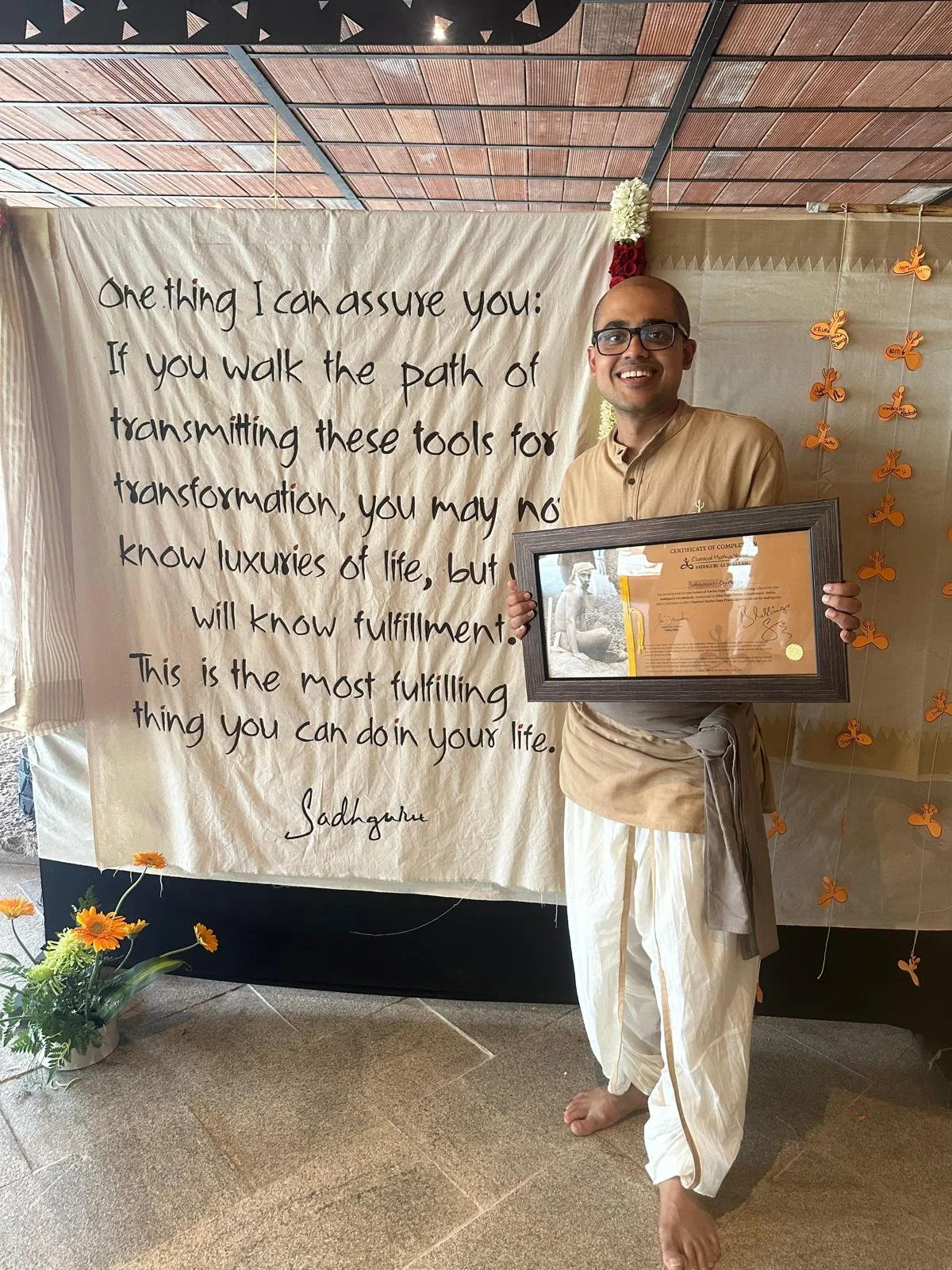 A man in traditional Indian attire holding a framed certificate, standing next to a large handwritten sign with an inspirational quote. The background includes orange butterfly decorations and a floral arrangement on the floor.