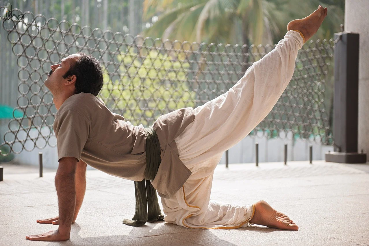 A man practicing yoga outdoors in a Cobra pose, wearing traditional white martial arts attire with a brown belt, on a concrete surface surrounded by a metal fence and tropical greenery.