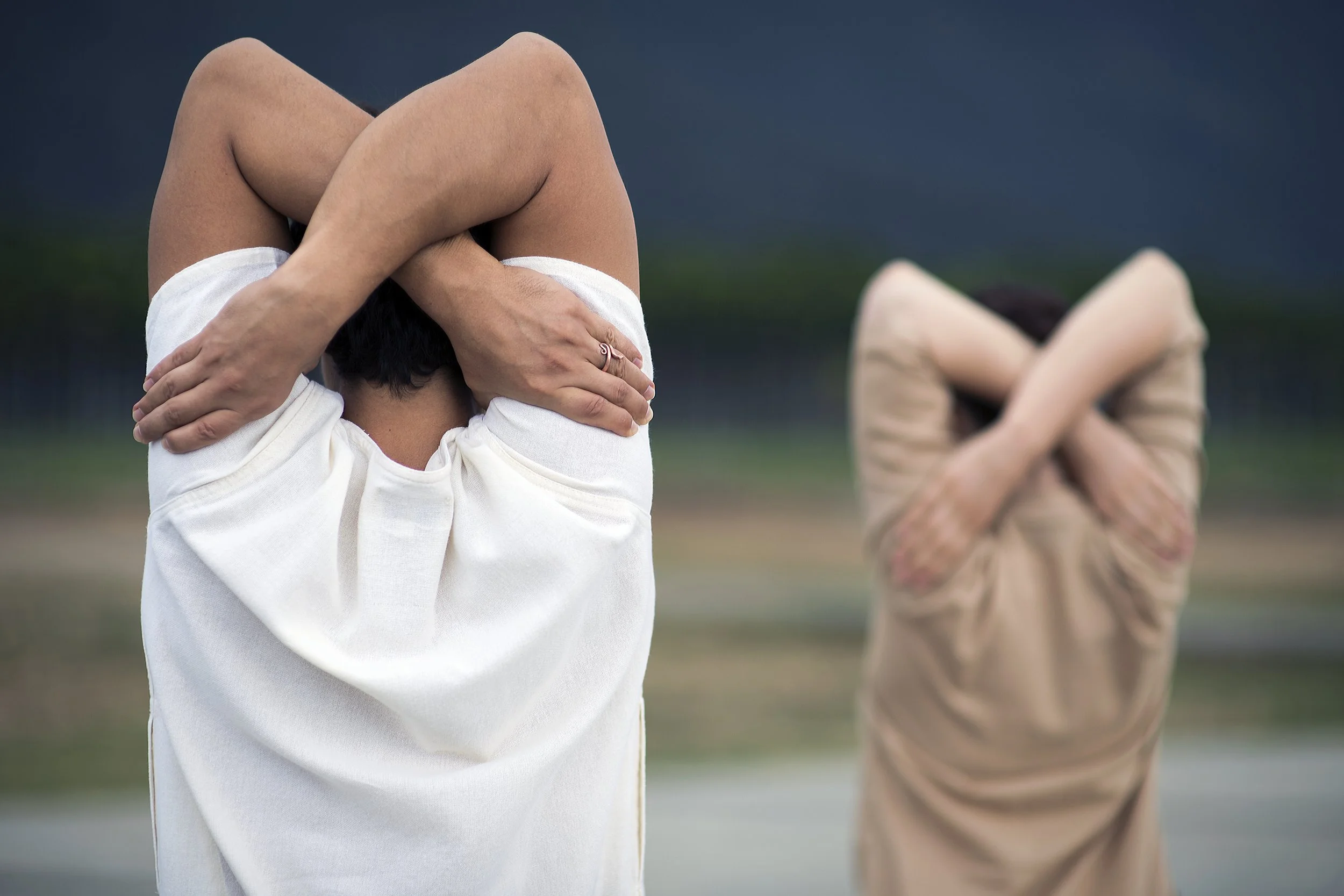 Two people dressed in loose clothing, standing outdoors, both stretching with arms crossed behind their heads. The background shows a cloudy sky and a landscape with trees.