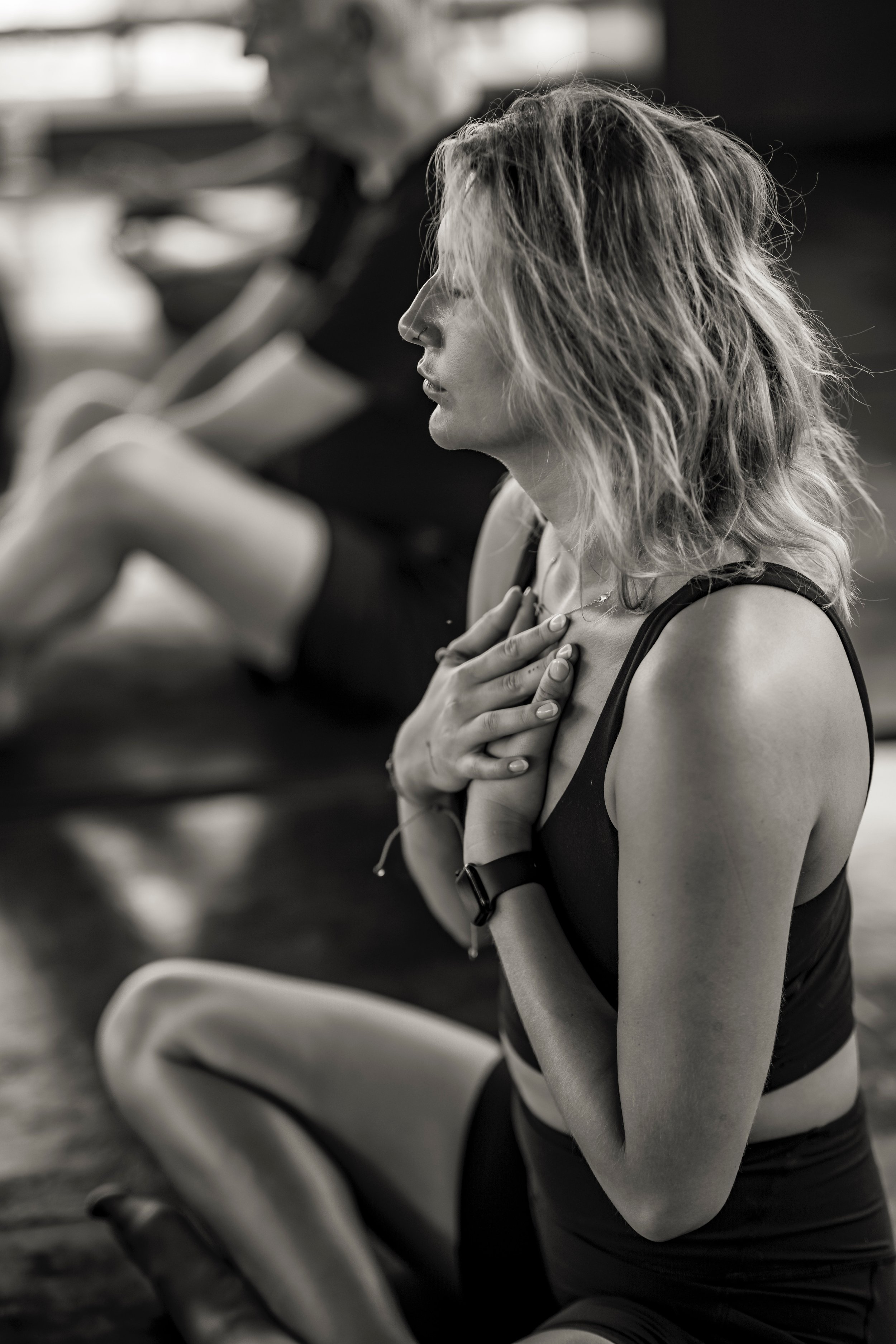 Black and white photo of a woman in a yoga pose with hands on her chest, eyes closed, and a man in the background also practicing yoga.