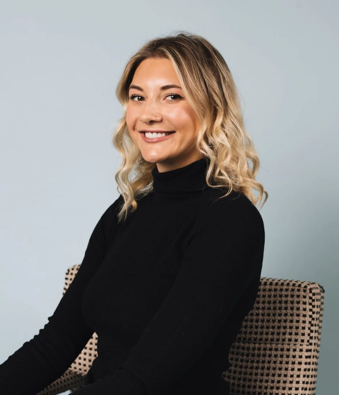 A young woman with blonde hair styled in loose waves, wearing a black turtleneck, smiling while sitting on a patterned chair against a plain light blue background.
