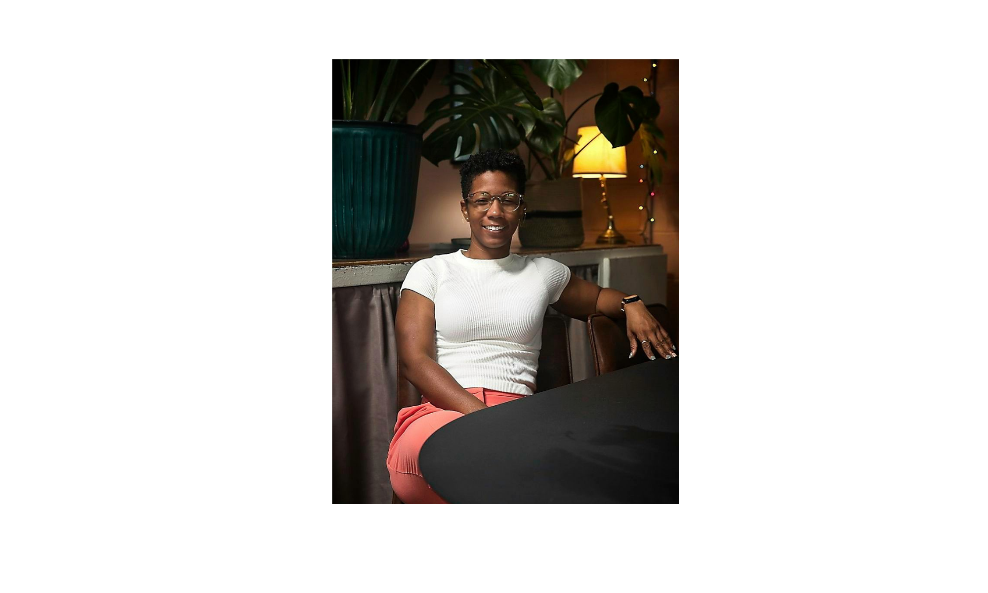 Woman with short curly hair, glasses, wearing a white top and pink pants, sitting at a table in a cozy, decorated room with large potted plant, warm lighting, and a lamp with string lights.