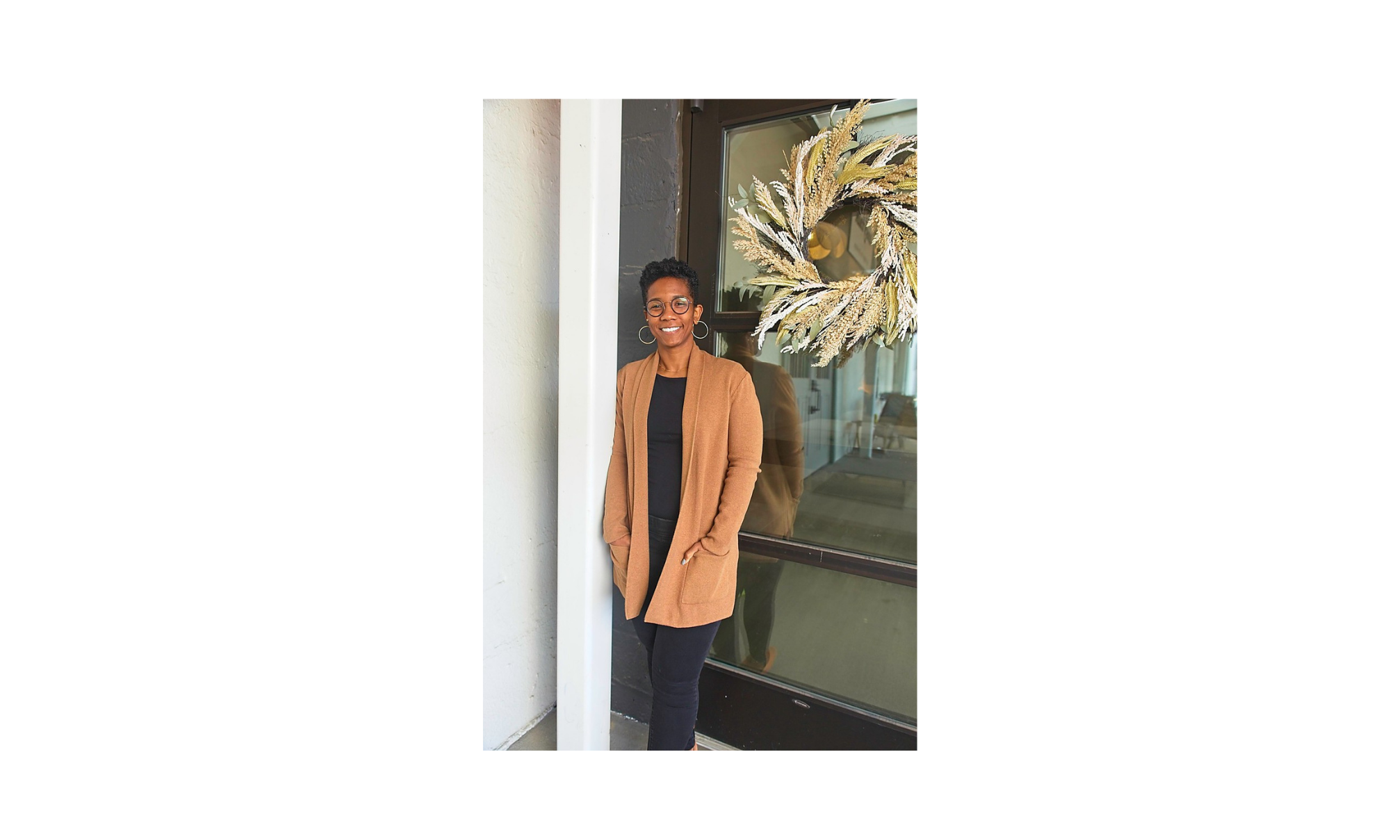 A woman stands outside near a glass window with a decorative wreath hanging on it, smiling at the camera.