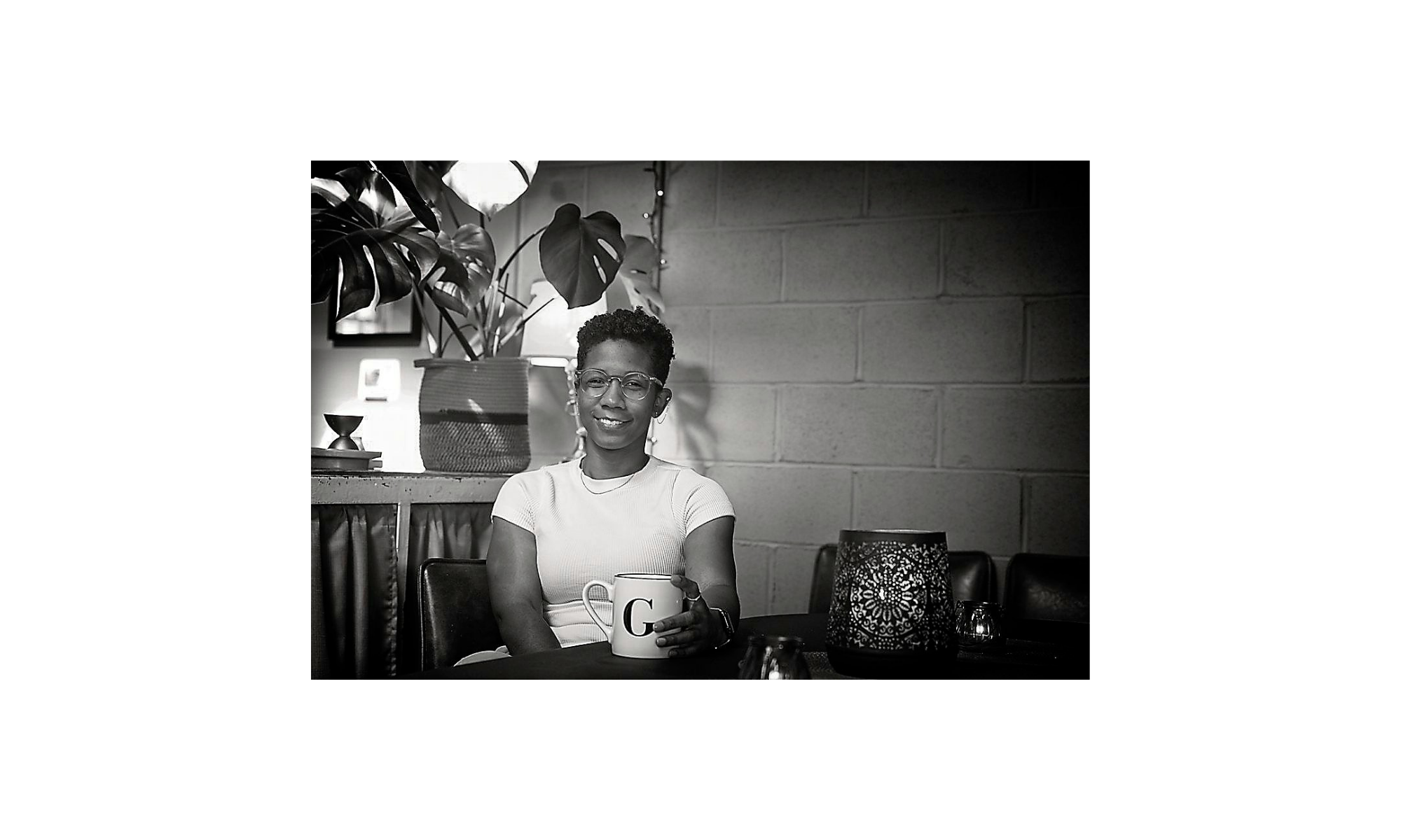 A woman with short hair and glasses smiling while holding a coffee mug with a letter G, sitting at a table with decorative candleholders, in a room with a brick wall and plants in the background.