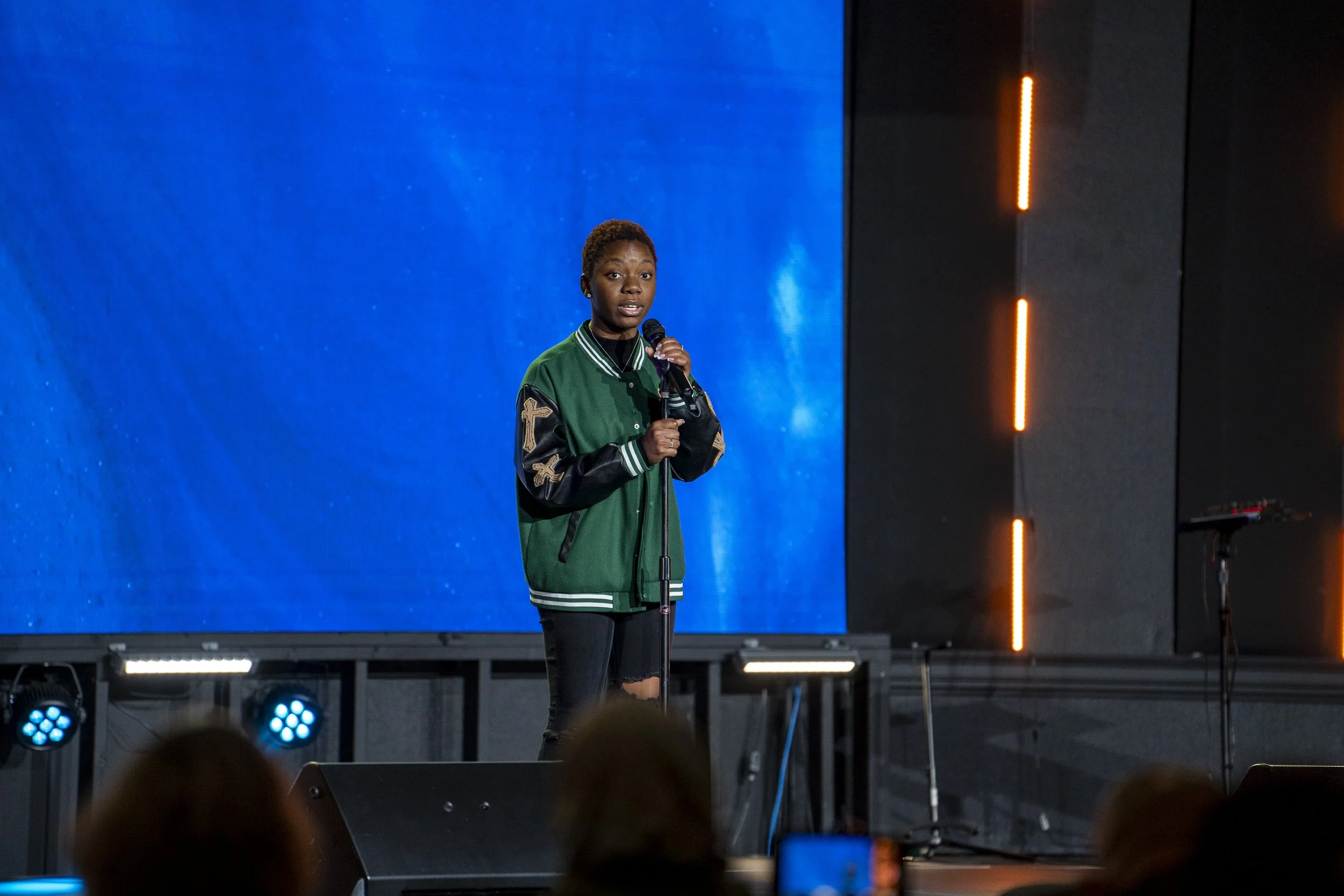 A young woman with short hair wearing a green and black varsity jacket with patches, standing on a stage with a blue background, holding a microphone and speaking to an audience.