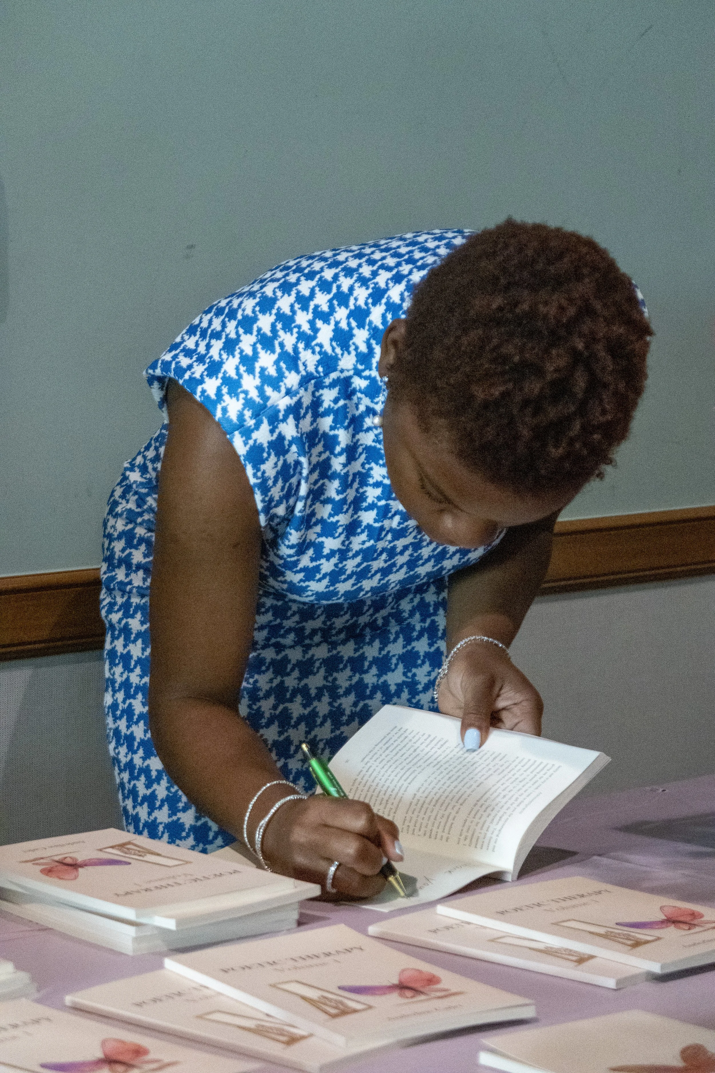 A woman with short curly hair wearing a blue and white houndstooth dress is signing a book at a table covered with several copies of the same book.