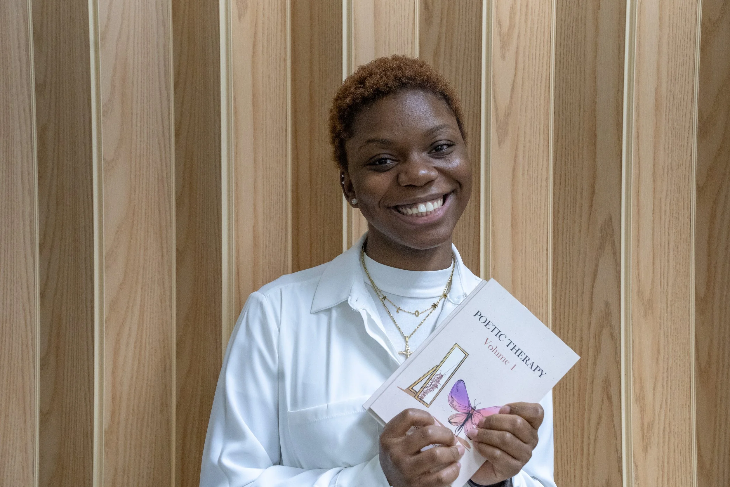 A woman with short curly hair, wearing a white shirt and layered necklaces, smiling and holding a booklet titled 'Poetic Therapy Volume 1' against a wood-paneled background.