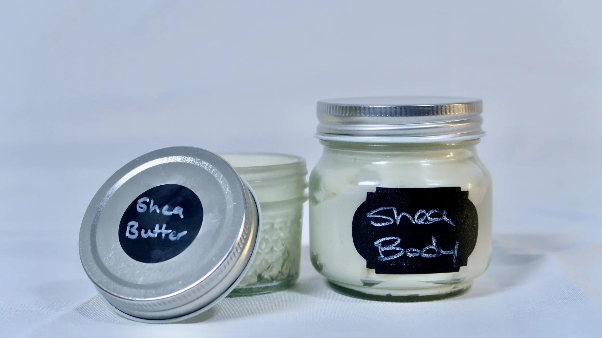 Two glass jars labeled 'Shea Butter' and 'Shea Body,' with the Shea Butter jar's lid closed and the Shea Body jar's lid slightly open, containing a creamy substance.