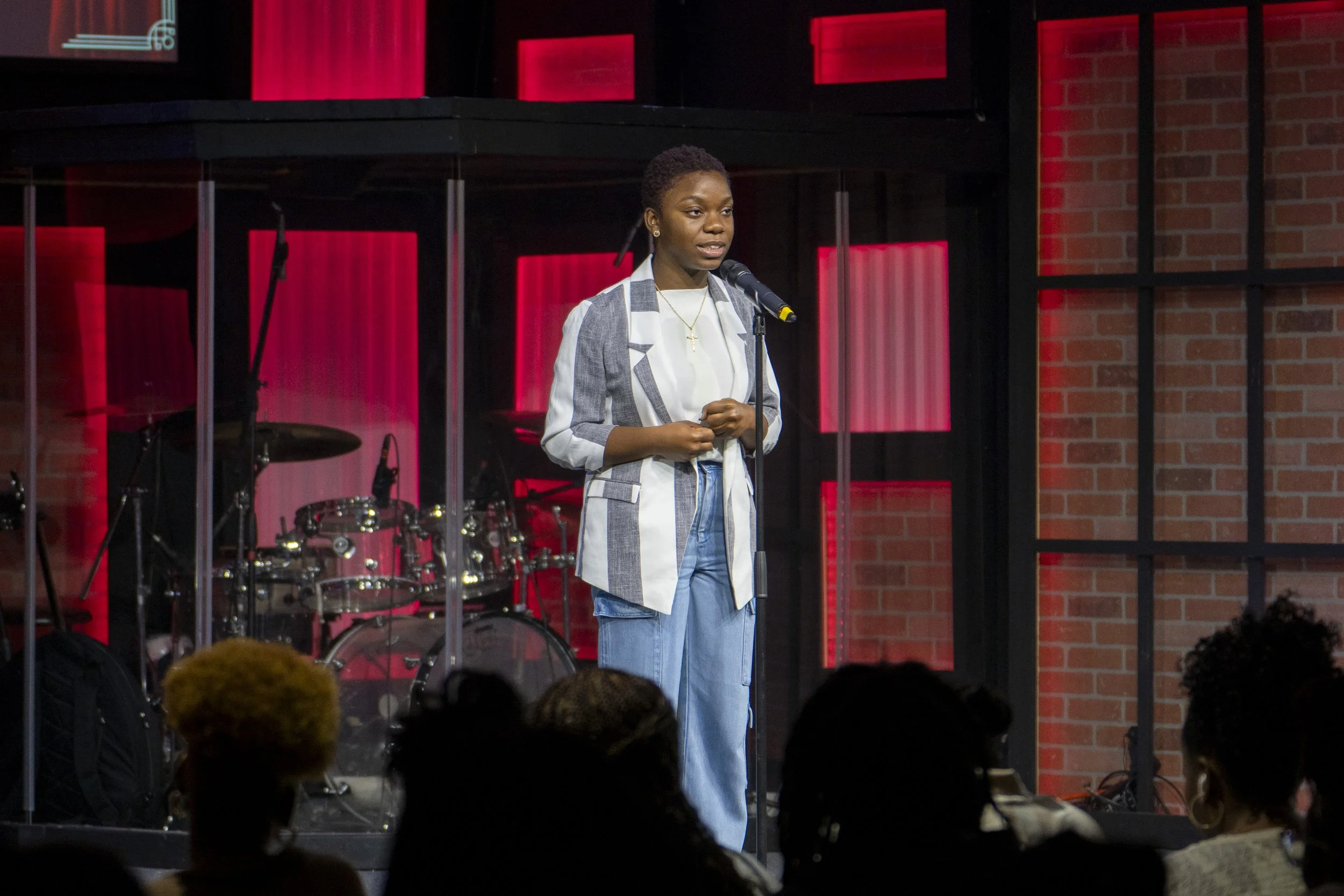 A young woman with short hair stands on stage, speaking into a microphone in front of a band set, with drums and guitars behind her, in a room with red illuminated windows and brick walls.