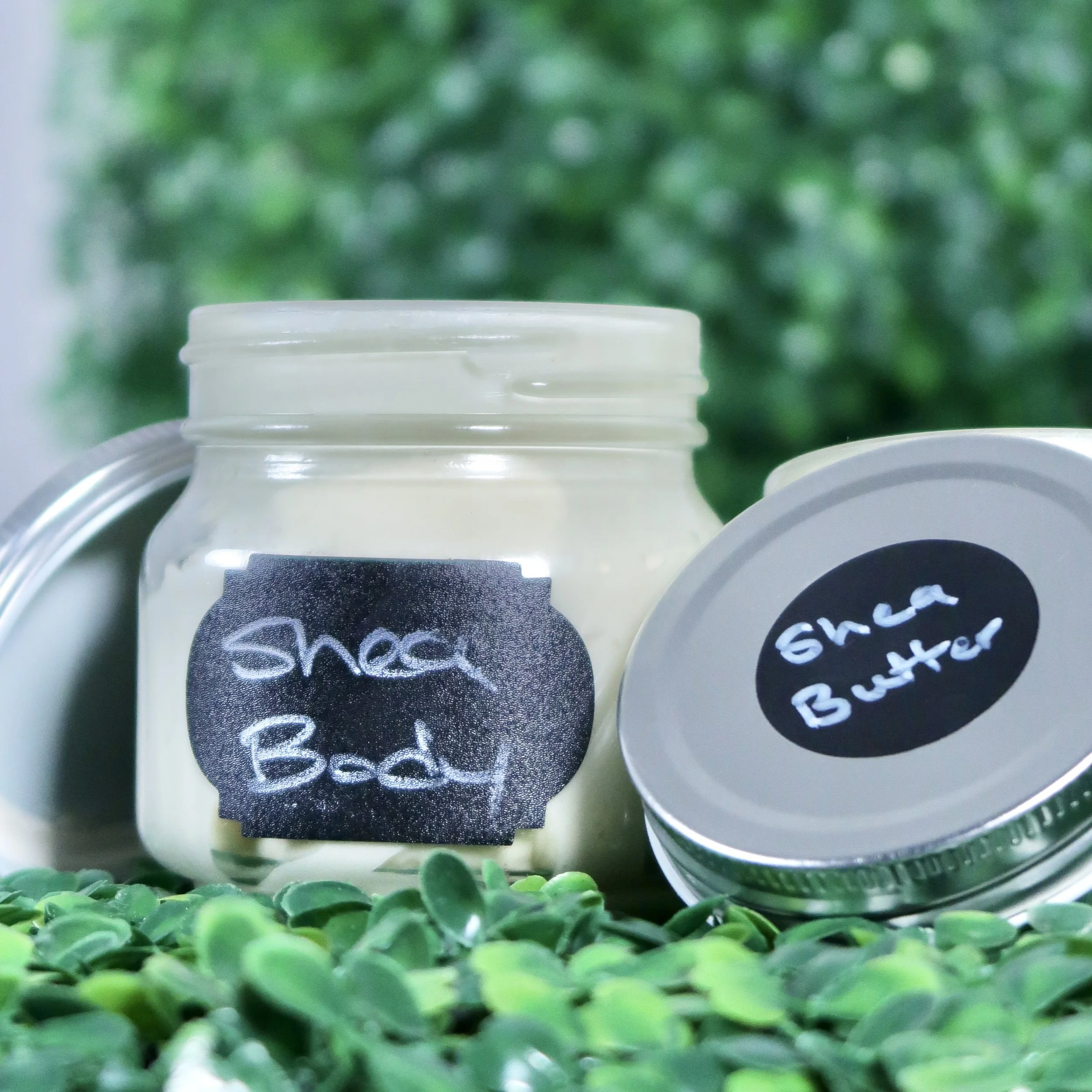 A jar labeled 'Shea Butter' and a tin labeled 'Ghee Butter' with green plants in the foreground and a blurred green background.