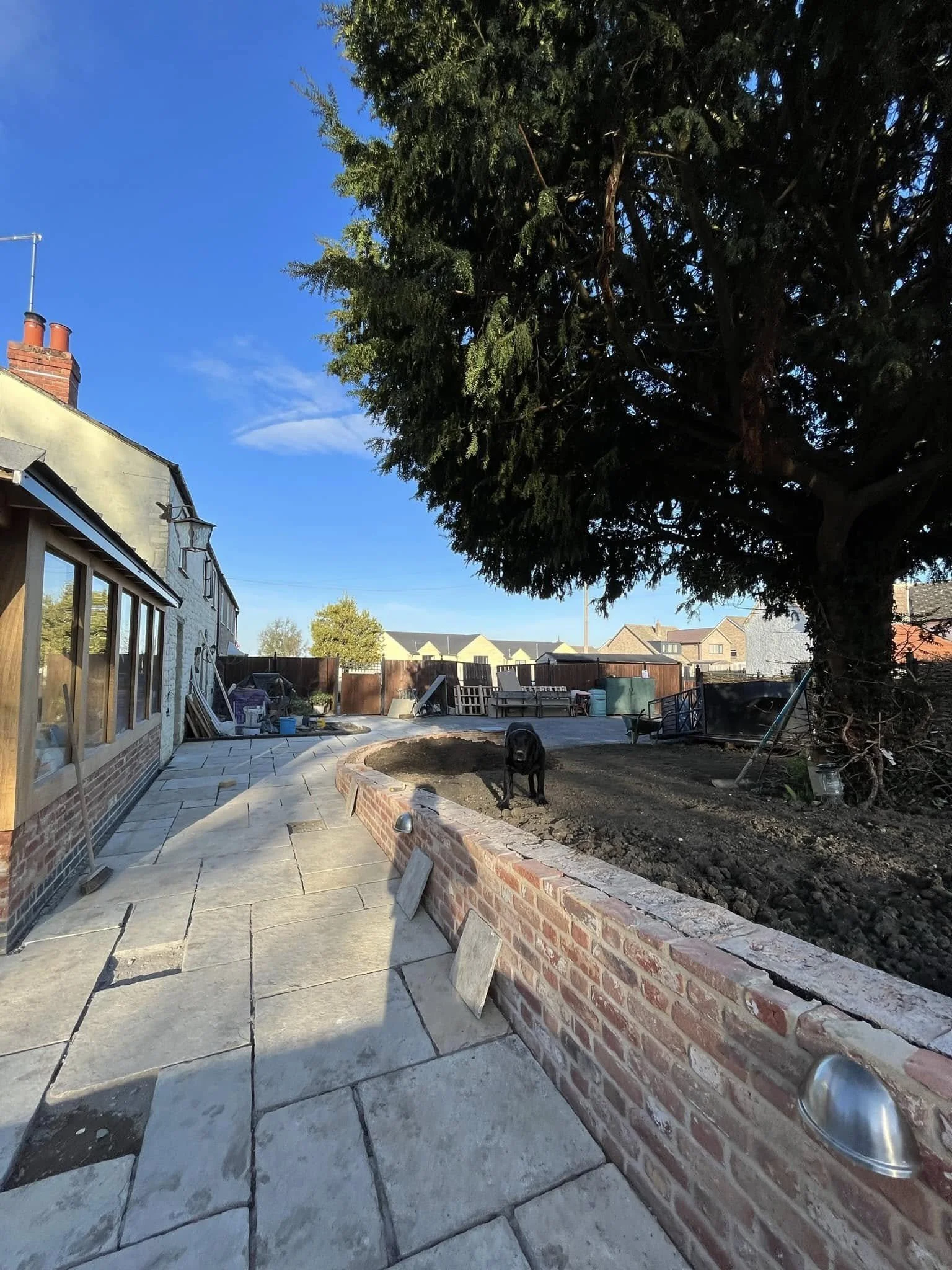 A backyard patio with a stone pathway, a large tree, and a dog standing near a garden bed. There's construction or gardening work underway, with building materials and tools visible in the background.