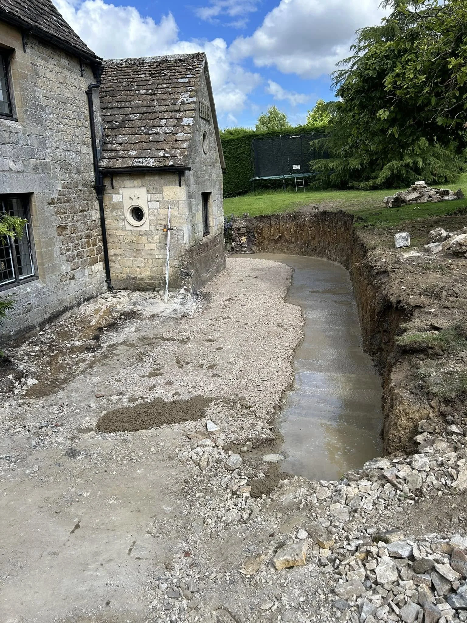 Construction site showing an excavated trench next to an old stone building with a measuring rod leaning against it, and a trampoline in the green backyard.