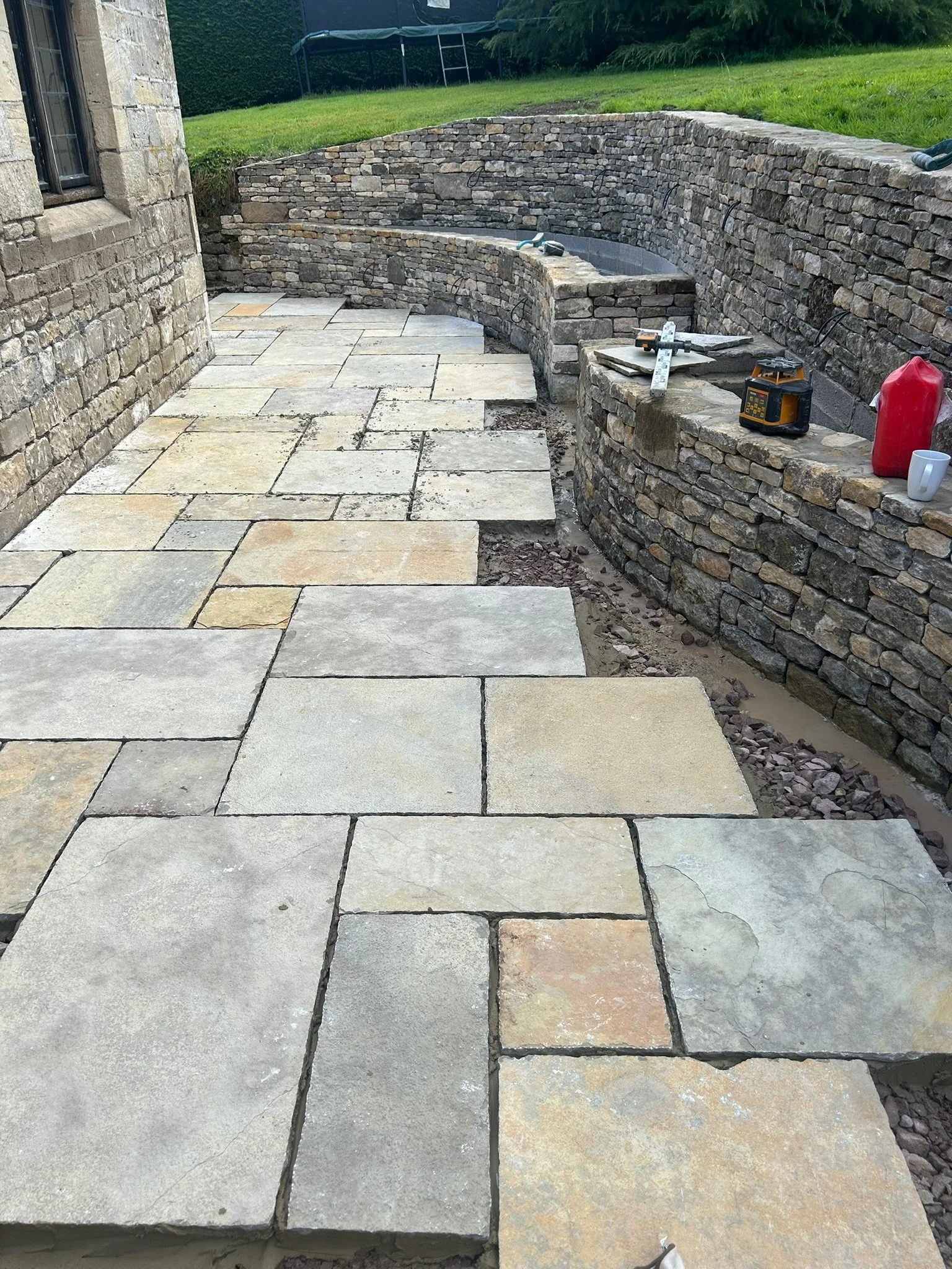 Outdoor patio under construction with large rectangular stone pavers laid in a pattern, and a curved stone wall built with stacked stones. Construction tools and materials are visible on the wall, with grassy yard and a trampoline in the background.