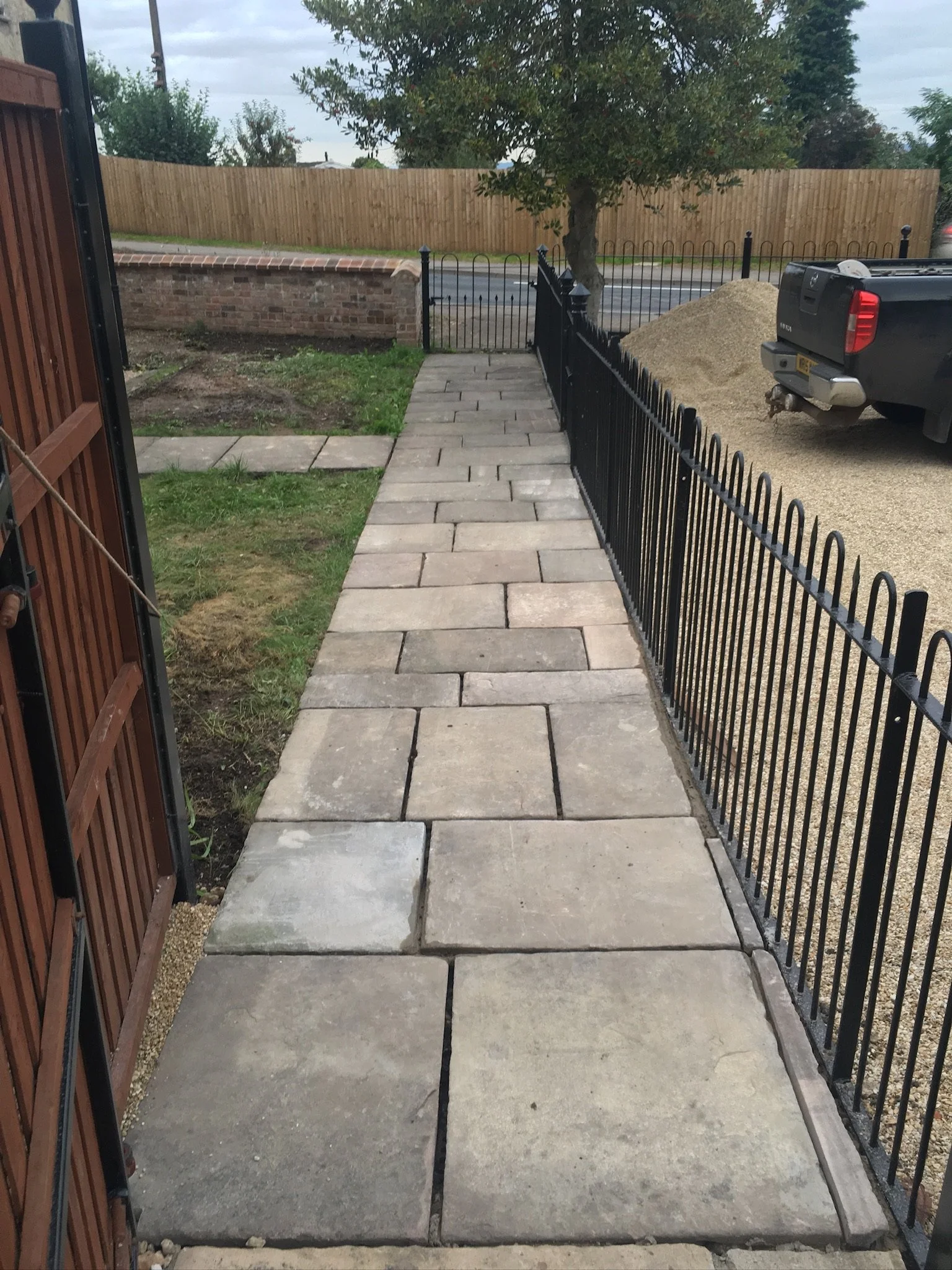 New stone sidewalk bordered by a metal fence on one side and a wooden fence on the other, with gravel and a parked truck in the background.